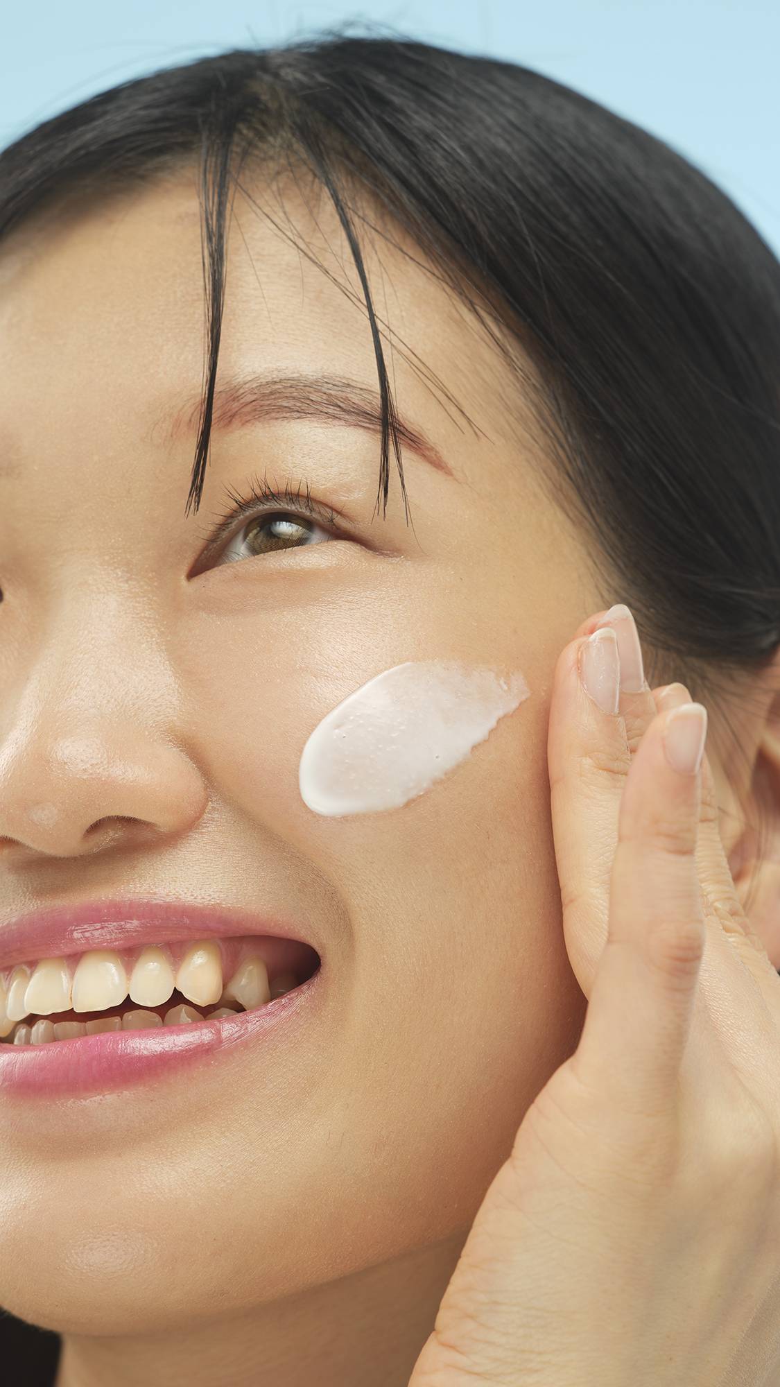 A close-up vertical shot of a smiling person with dark hair, applying a creamy white swatch of Calm primer to their cheek with a finger. The product is being blended into their skin, set against a bright blue background.