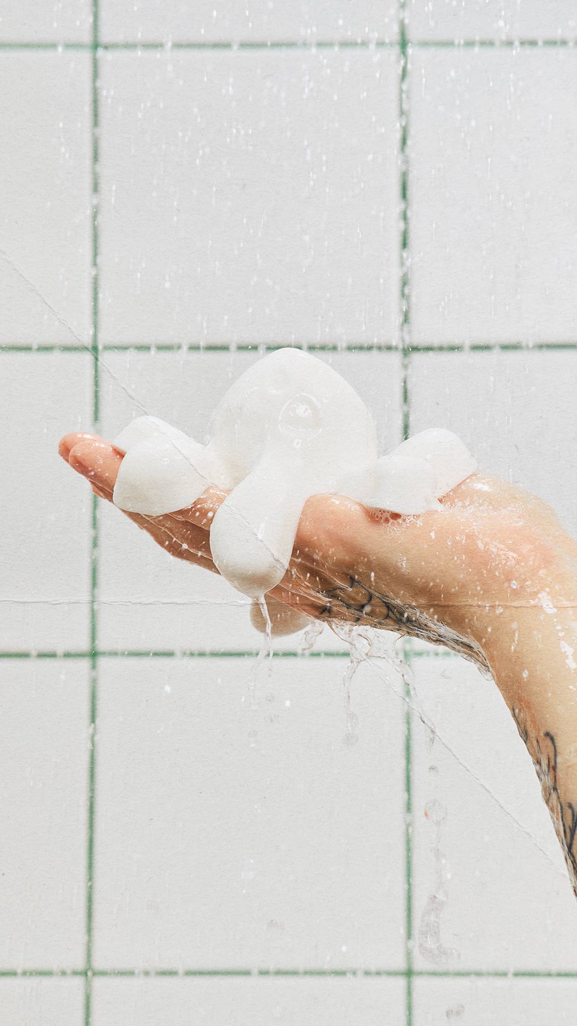 A tattooed hand holds a jiggly, white ghost-shaped shower jelly under streaming water in a shower. Water splashes off the product against a background of white square tiles with green grout.