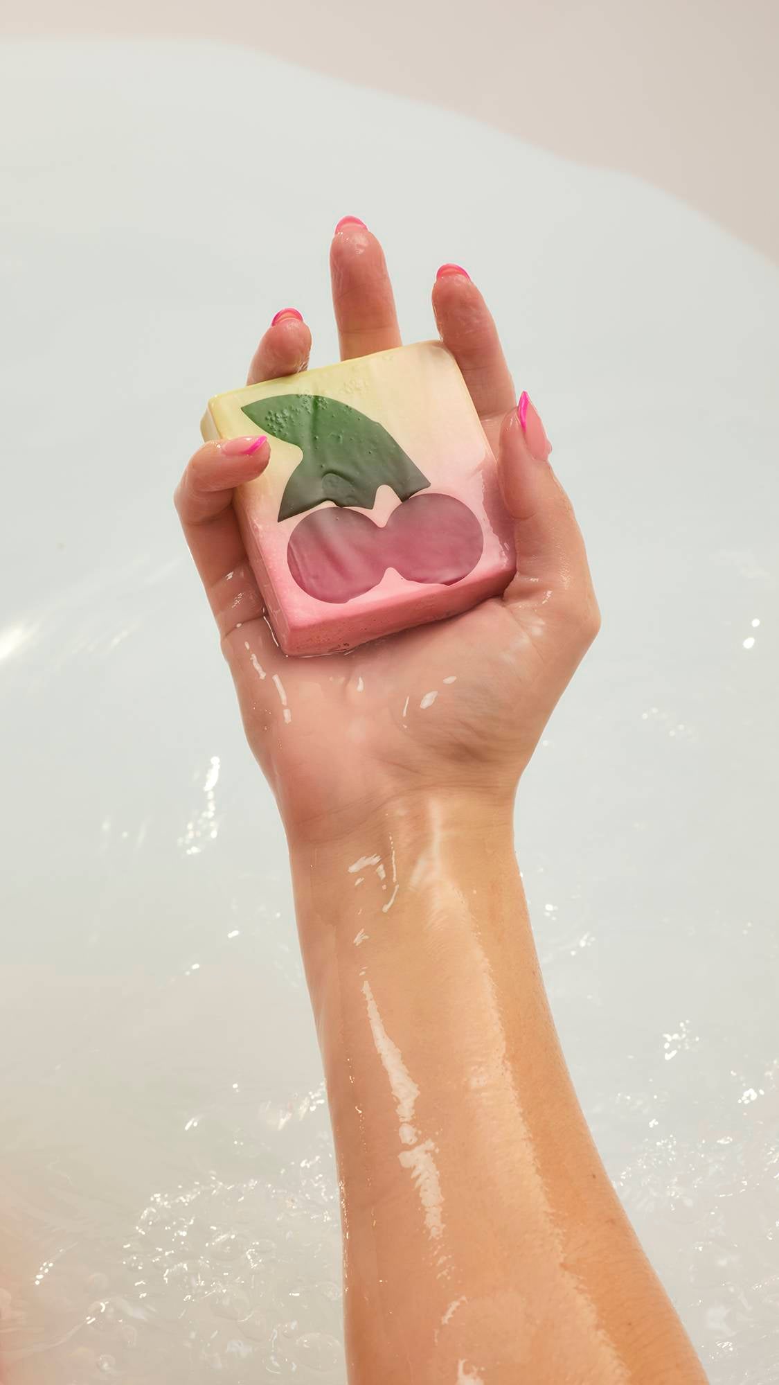 A person with pink-tipped nails holds the cream-coloured Cherry On Top soap bar over a white bathtub. The soap features a large dark pink cherry and green leaf design, its surface wet from the bathwater.