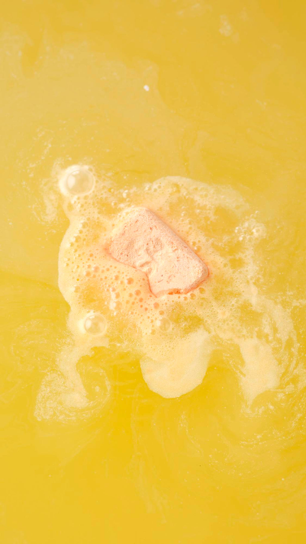 An aerial view of the Cold Water Soother bath bomb dissolving. The product creates a thick layer of white velvety foam that ripples and swirls across the surface of yellow water.