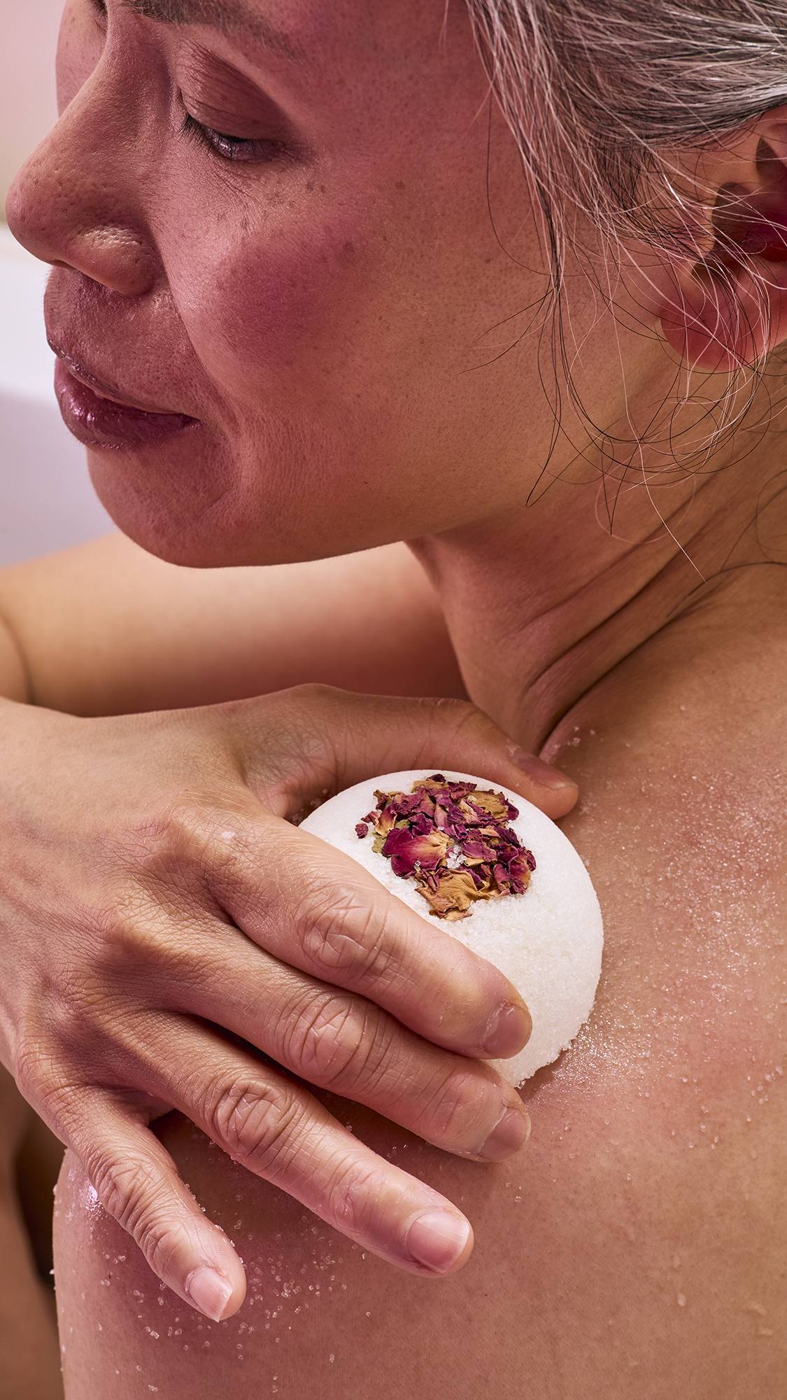 A model in a bath uses a white, dome-shaped "Comforter Rose" solid body scrub. They are rubbing the textured surface against their shoulder, leaving a fine, white exfoliating residue on their skin.