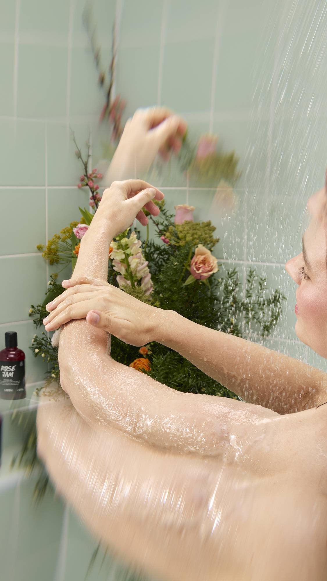 A person stands under a shower stream against a background of mint green tiles and flowers, massaging Crème Brûlée Body Scrub into their wet arm. A bottle of "Rose Jam" Shower Gel is visible on the shelf in the background.