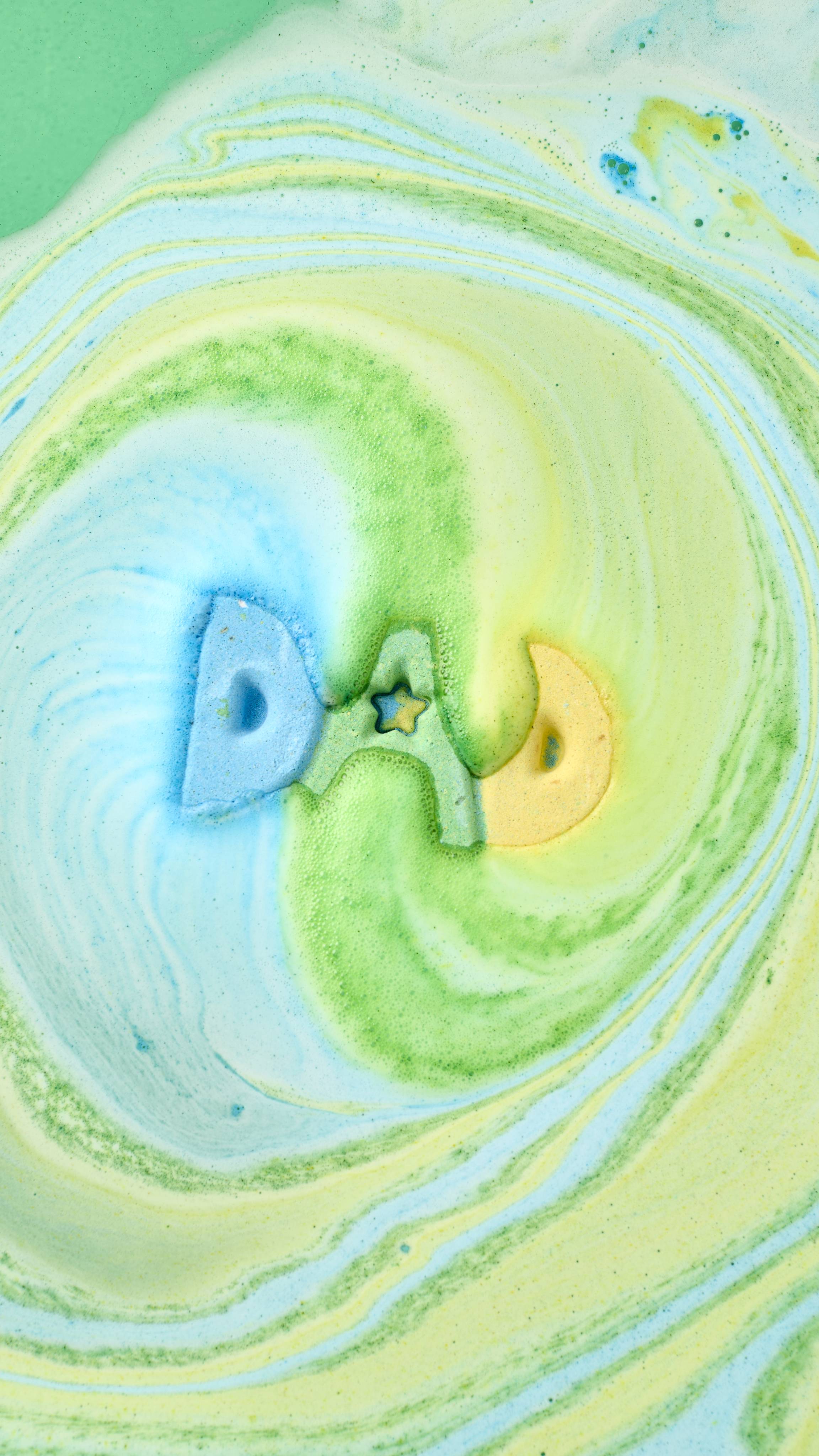 An aerial view of the "DAD" bath bomb dissolving in a bathtub. The blue, green, and yellow letters create a hypnotic swirl of colourful foam on the water's surface, forming vibrant marbled patterns of teal, lime, and soft yellow.
