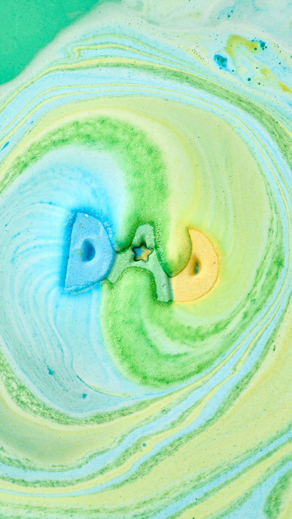 An aerial view of the "DAD" bath bomb dissolving in a bathtub. The blue, green, and yellow letters create a hypnotic swirl of colourful foam on the water's surface, forming vibrant marbled patterns of teal, lime, and soft yellow.