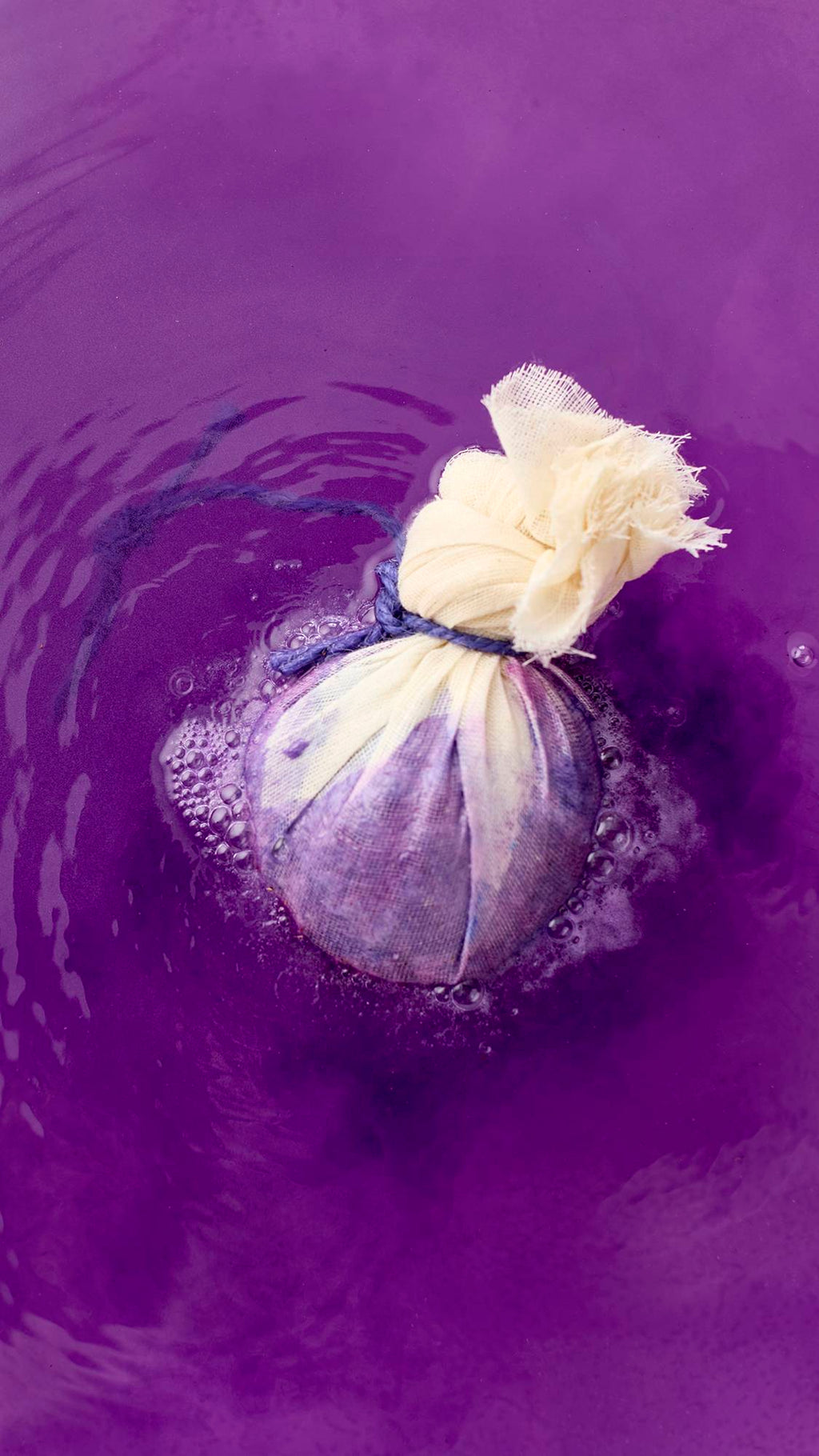 The spherical bath bomb is wrapped in cream-colored muslin, tied with a purple cord, and half-submerged in water. The water reflects the vibrant purple, with ripples spreading across the surface.