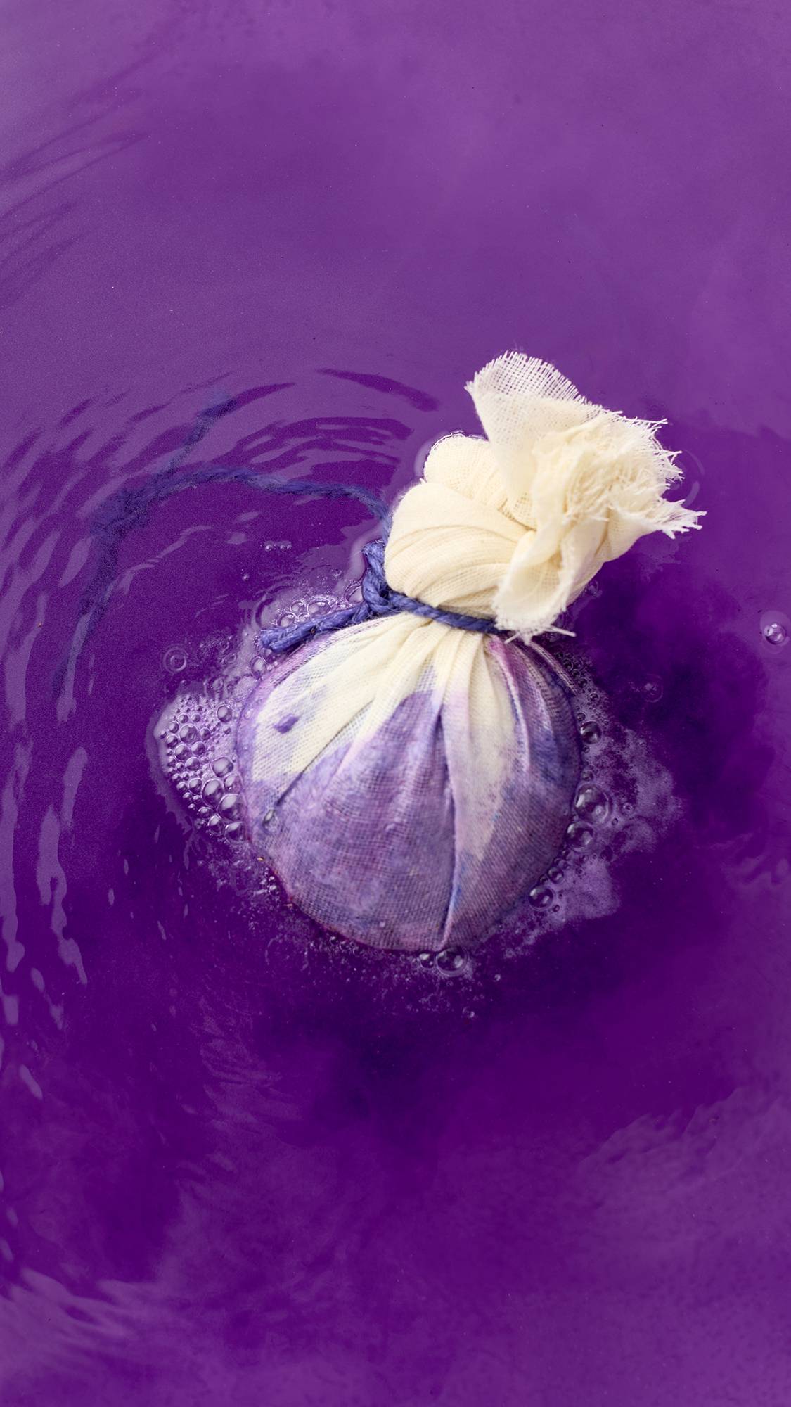 The spherical bath bomb is wrapped in cream-colored muslin, tied with a purple cord, and half-submerged in water. The water reflects the vibrant purple, with ripples spreading across the surface.
