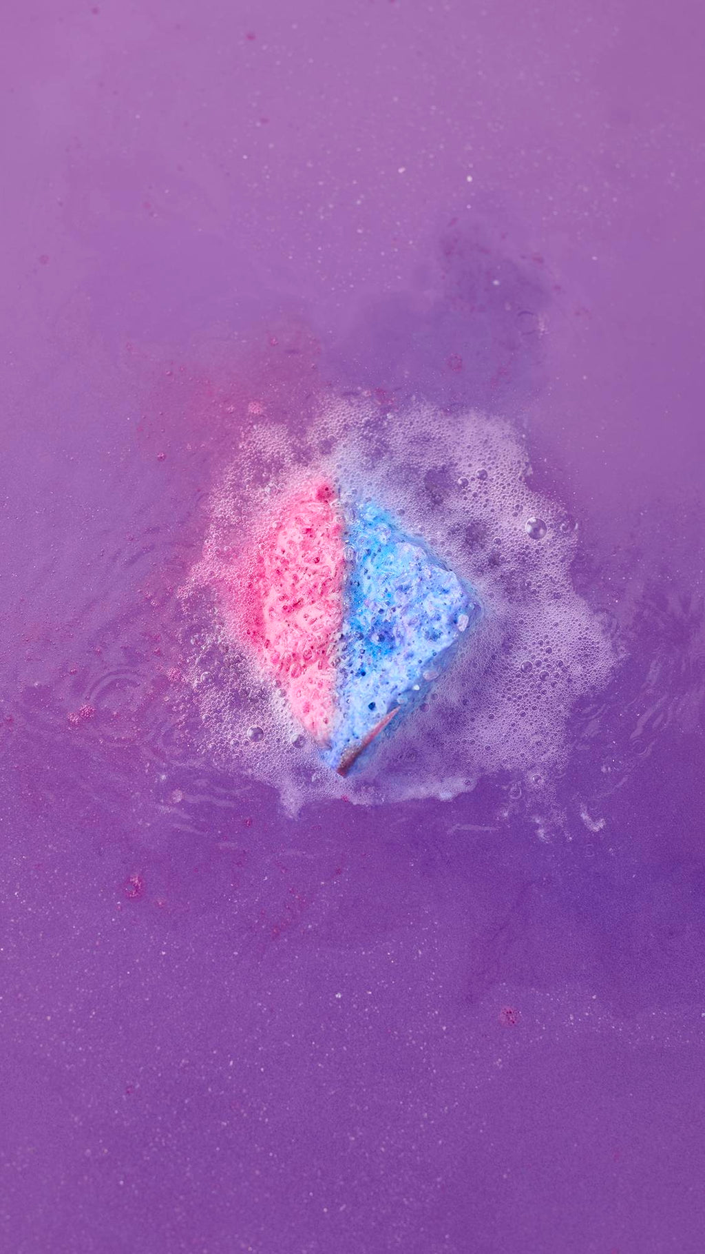 A top-down view of the Deep sleep Salt Cube bath bomb fizzing in deep purple water. The pink and blue cube is surrounded by a ring of frothy white foam and bubbles as it dissolves.