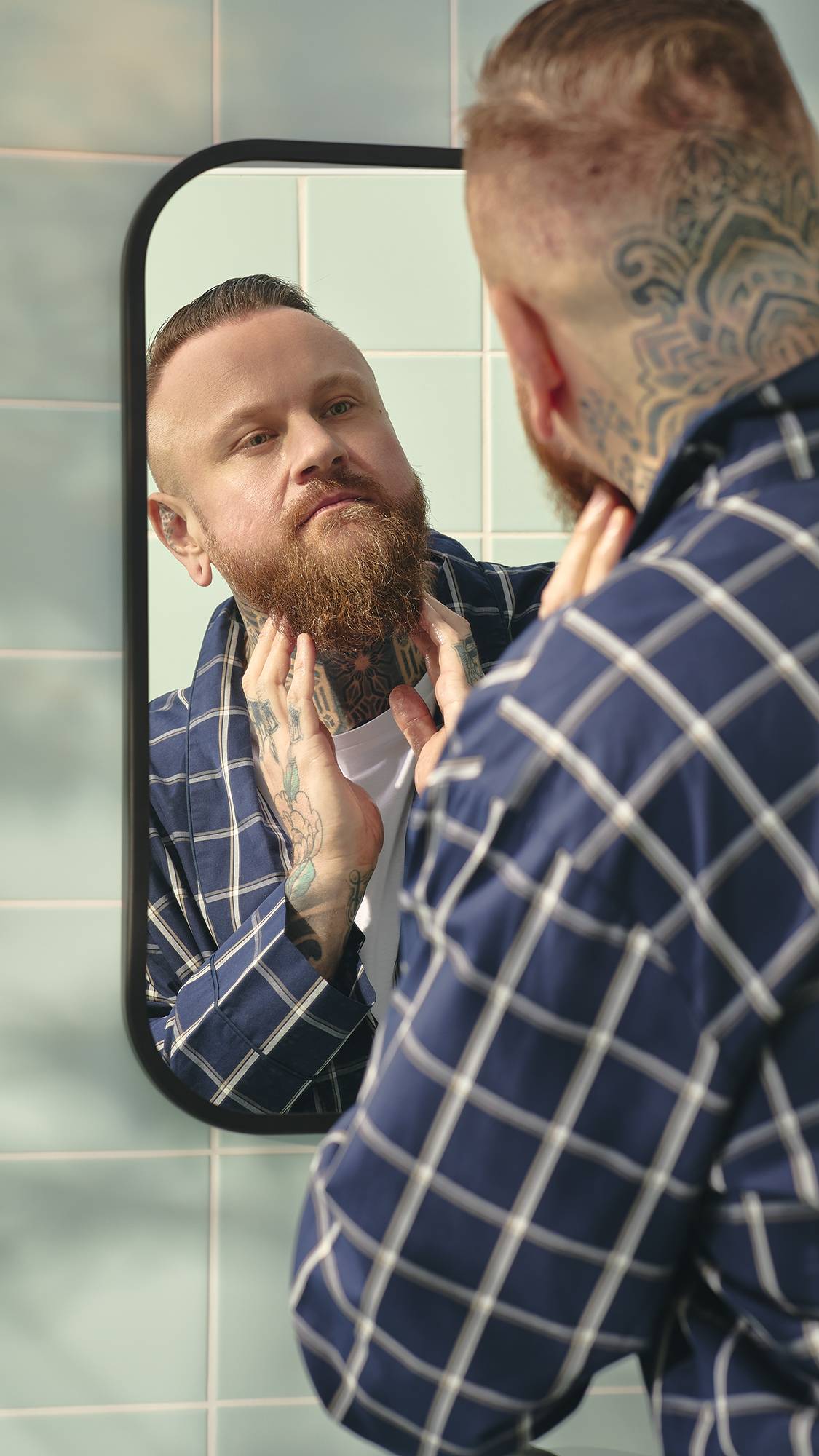 A model with a ginger beard and a blue-and-white checked dressing gown looks into a bathroom mirror. They have extensive tattoos on their neck and hands, and are using their fingers to massage the "Dirty" Hair and beard oil in.