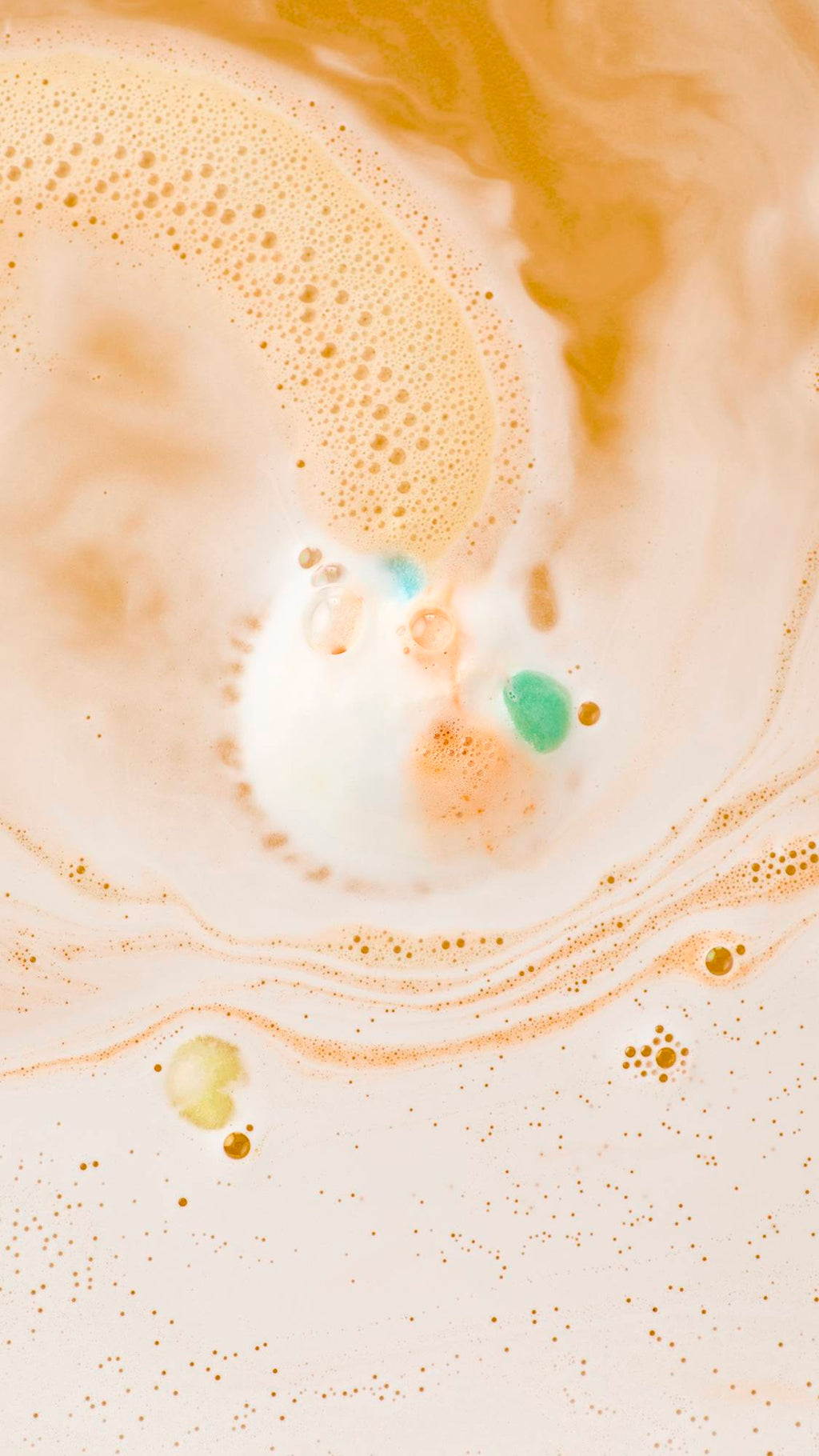 An aerial view of a "Dragon'/s Egg" bath bomb dissolving in orange-tinted water. Bright green, blue, and orange foam swirls outwards, mixing with white frothy bubbles to create a marbled, abstract pattern across the surface.
