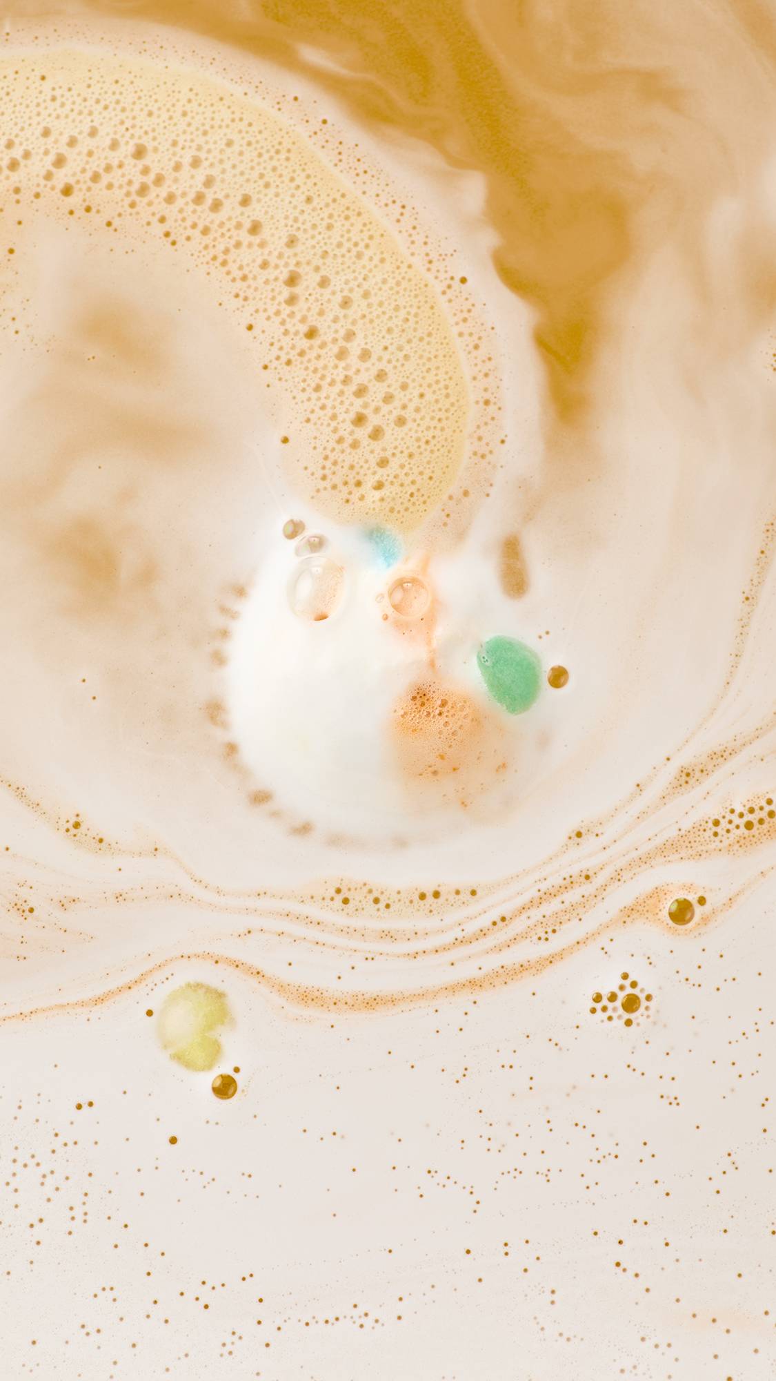 An aerial view of a "Dragon'/s Egg" bath bomb dissolving in orange-tinted water. Bright green, blue, and orange foam swirls outwards, mixing with white frothy bubbles to create a marbled, abstract pattern across the surface.