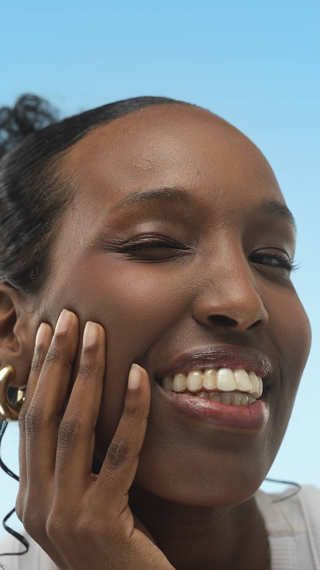 A person with dark skin smiles widely, touching their cheek. They have a subtle shimmer and gold hoop earrings. The smooth finish on their skin suggests the Emotional Brilliance ~ Finishing Powder, against a bright blue background.