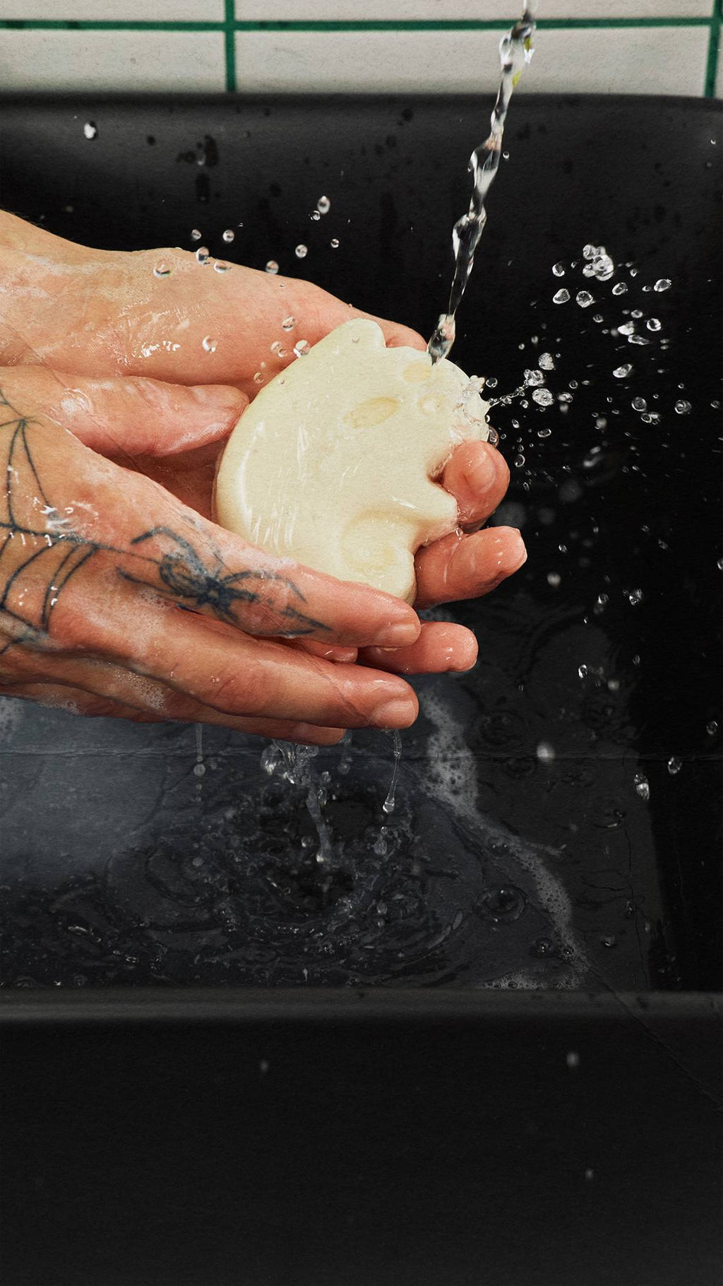 Tattooed hands hold an off-white, ghost-shaped bar of soap under running tap water over a black basin. Water splashes as the soap is lathered. White wall tiles with green grout lines are visible at the top.