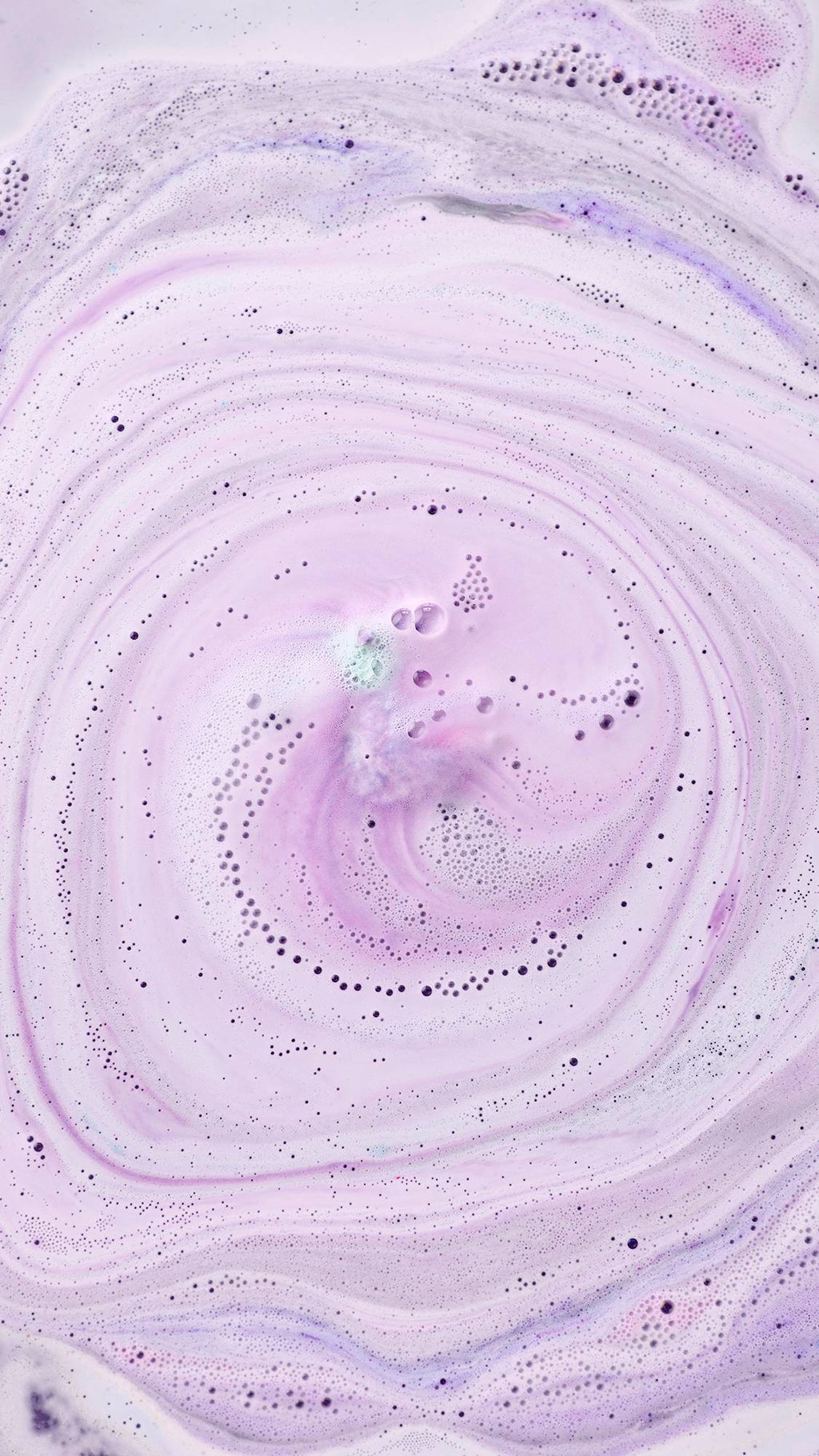 An aerial view of pale lilac bathwater covered in swirling patterns of white and purple foam. Tiny bubbles and darker purple droplets create intricate trails across the surface, spiralling towards the fizzing "Goddess" bath bomb.