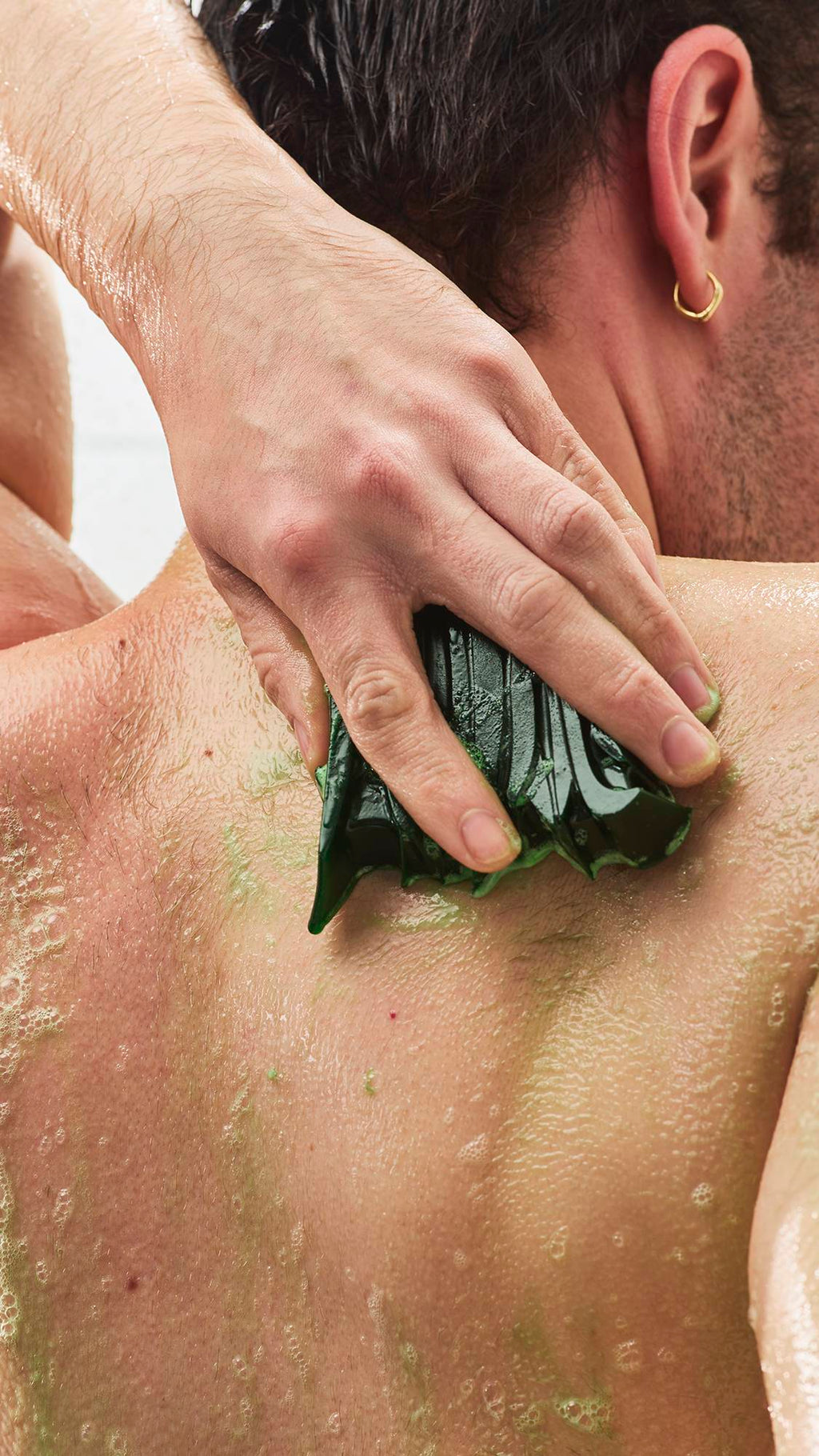 A close-up view from behind of a person rubbing a textured, dark green shower gummy soap across their wet back, creating a light green lather with soapy bubbles.