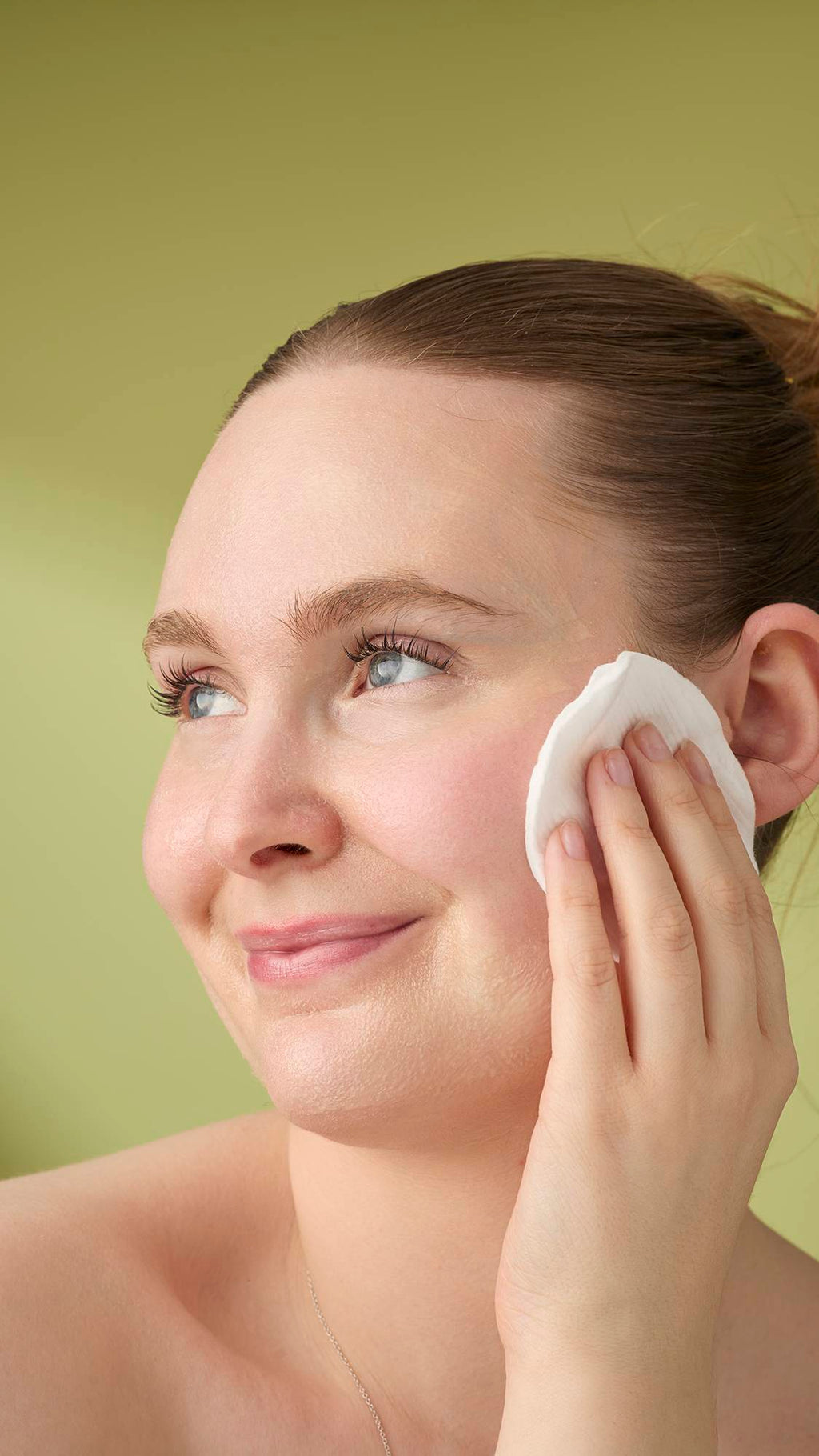 A model with light hair smiles while wiping the yellow Honey, Beeswax And Almond Oil cleanser with a cotton pad. She is shown from the shoulders up against a solid olive-green background, highlighting the creamy texture of the facial product.