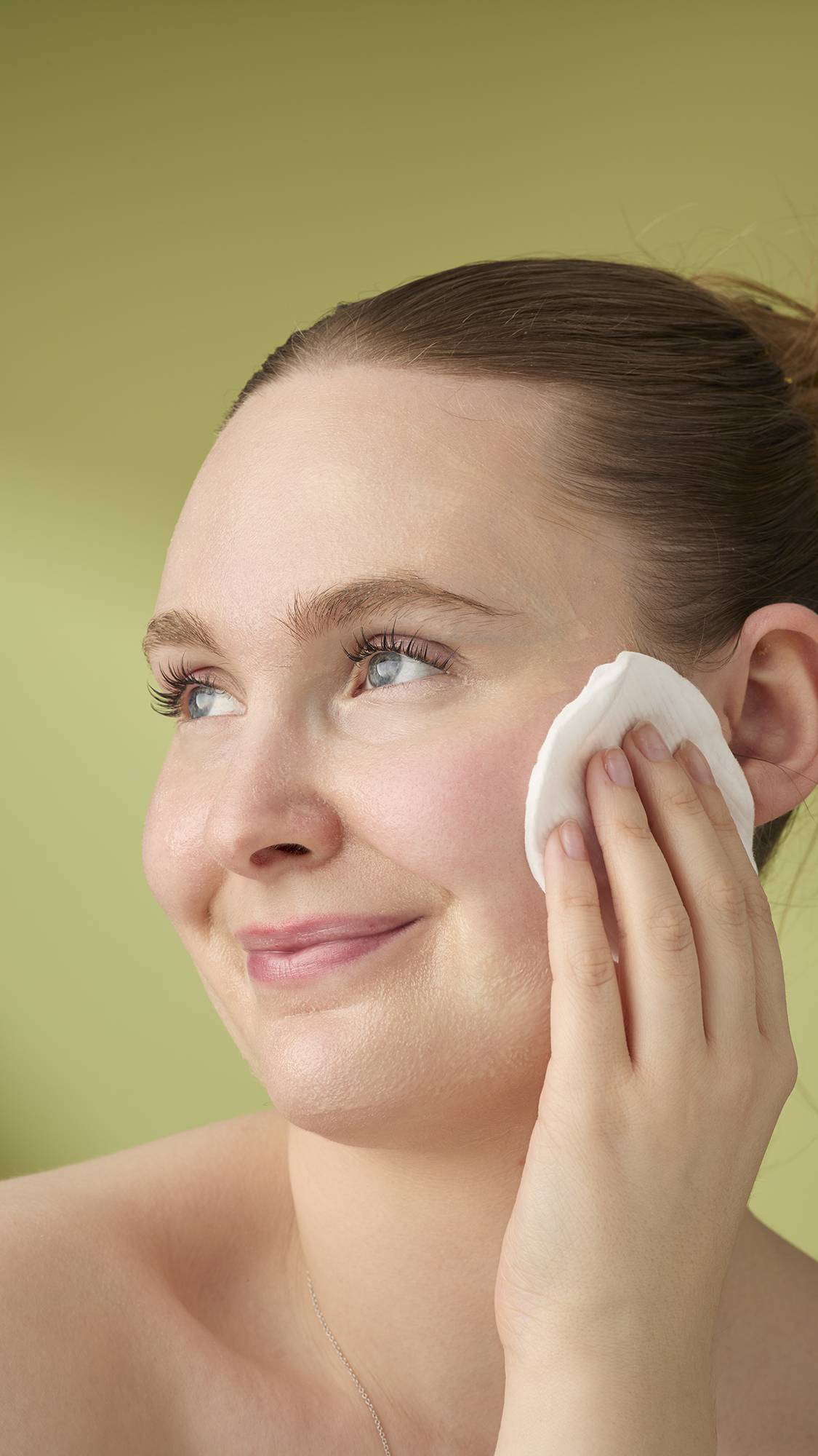 A model with light hair smiles while wiping the yellow Honey, Beeswax And Almond Oil cleanser with a cotton pad. She is shown from the shoulders up against a solid olive-green background, highlighting the creamy texture of the facial product.