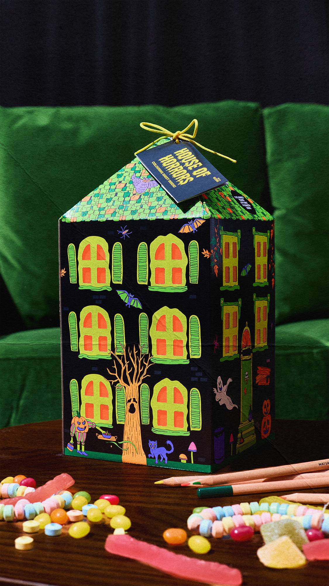 A Lush 'House of Horrors' gift box shaped like a haunted house sits on a wooden table. A green velvet couch is in the background. In the foreground, colourful sweets and coloured pencils are scattered across the table.