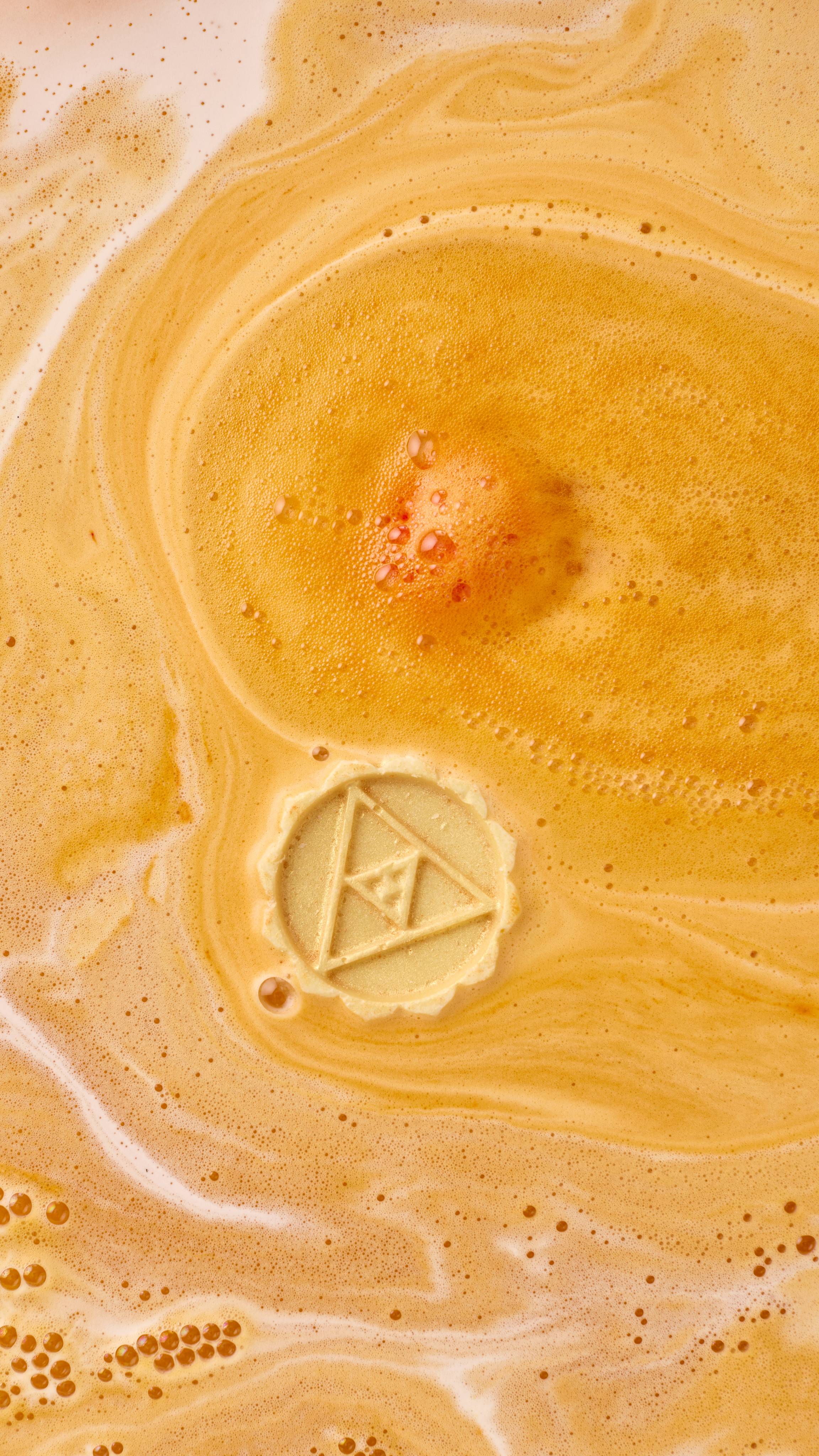 An aerial view of the bathwater after using the Karma macaron-shaped bath bomb. The orange product has dissolved, leaving spiralling swirls of thick, velvety, golden foam. The solid oil central layer floats, slowly dissolving.