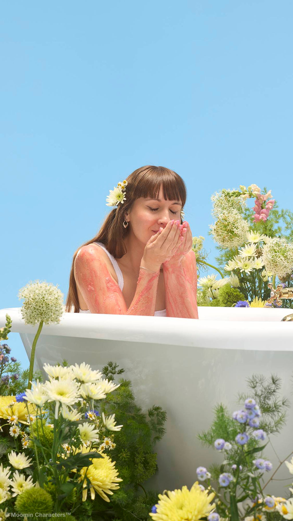 A close-up shot of a person with flowers in their hair sitting in a bathtub, bringing their hands to their nose to smell a pred "Snufkin" soap, which is lathered on their arms. Yellow and white wildflowers surround the tub against a clear blue sky.