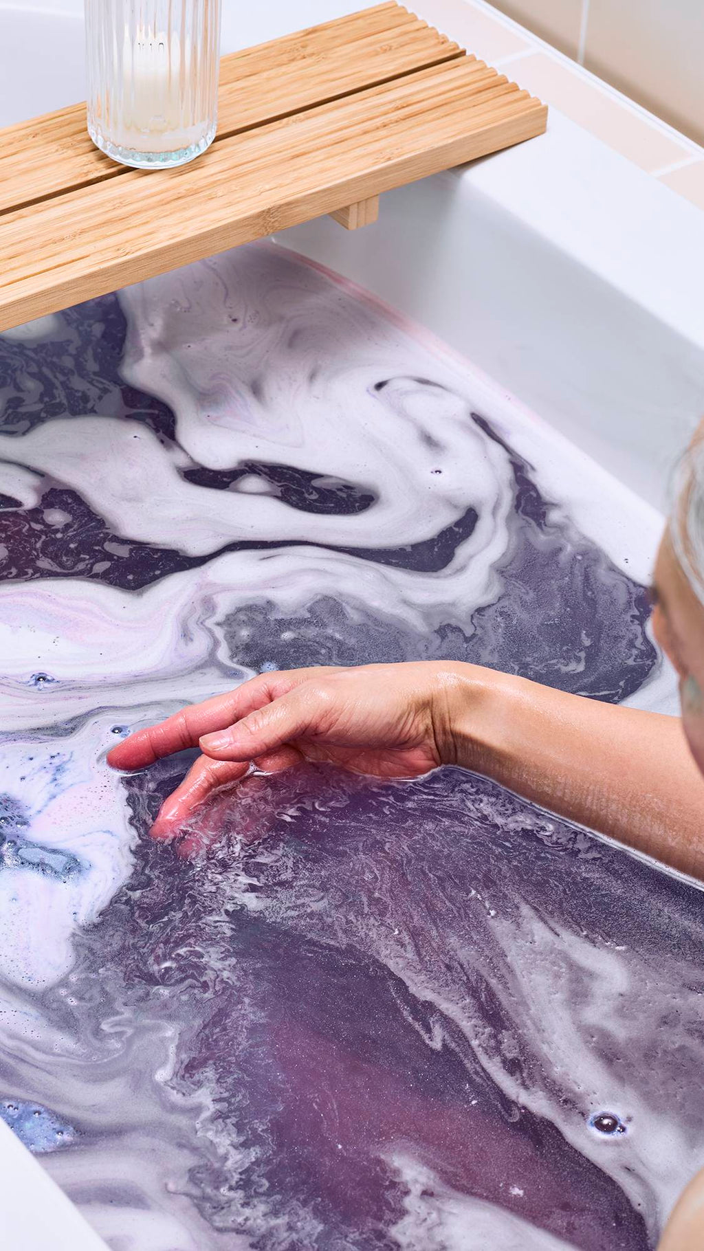 An angled aerial view of the model in the bath after using the "Magik Mushroom" bath bomb. The water has turned a deep purple colour, and there is a shimmering, sparkling, silver lustre throughout as the model moves their arm across the surface.