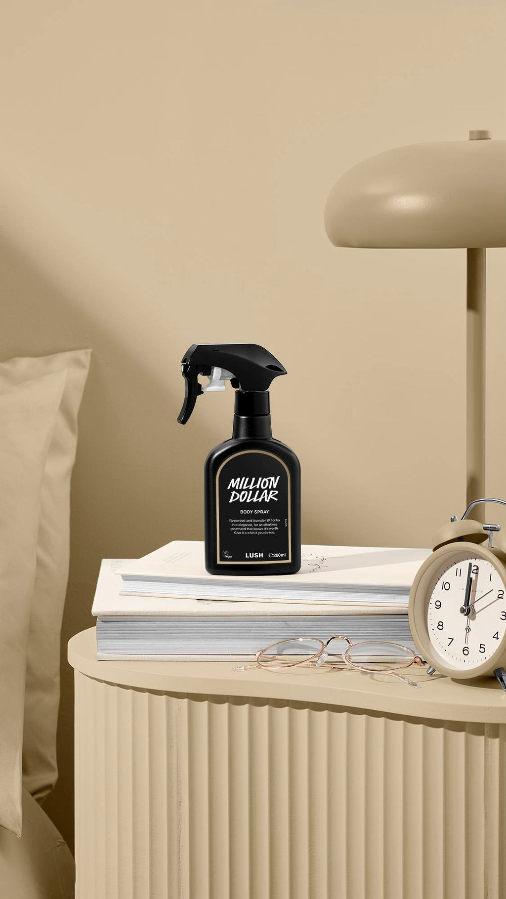 A black bottle of Lush 'Million Dollar' body spray stands on a stack of books on a beige bedside table. A matching lamp, an alarm clock, and a pair of glasses are also on the table against a neutral wall and pillow.