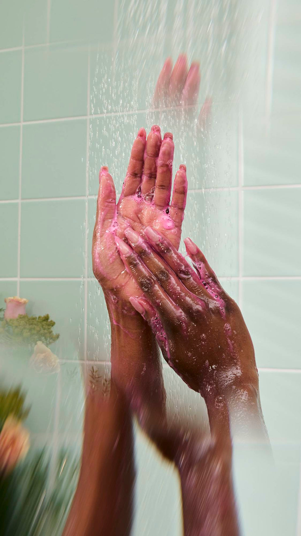Wet hands lather a pink, soapy foam under a stream of shower water. The scene highlights the colorful suds created by the product against a backdrop of mint green tiles and blurred flowers.
