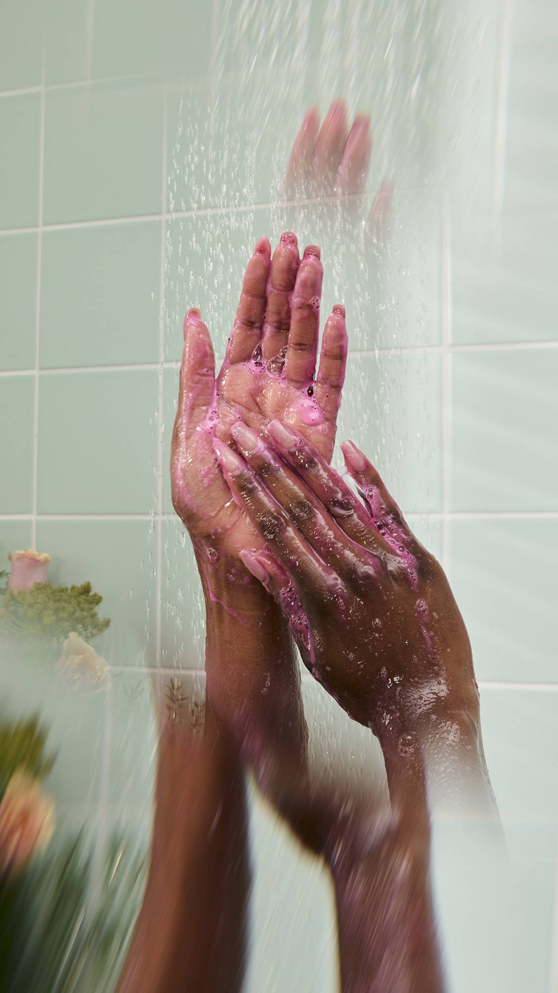 Wet hands lather a pink, soapy foam under a stream of shower water. The scene highlights the colorful suds created by the product against a backdrop of mint green tiles and blurred flowers.