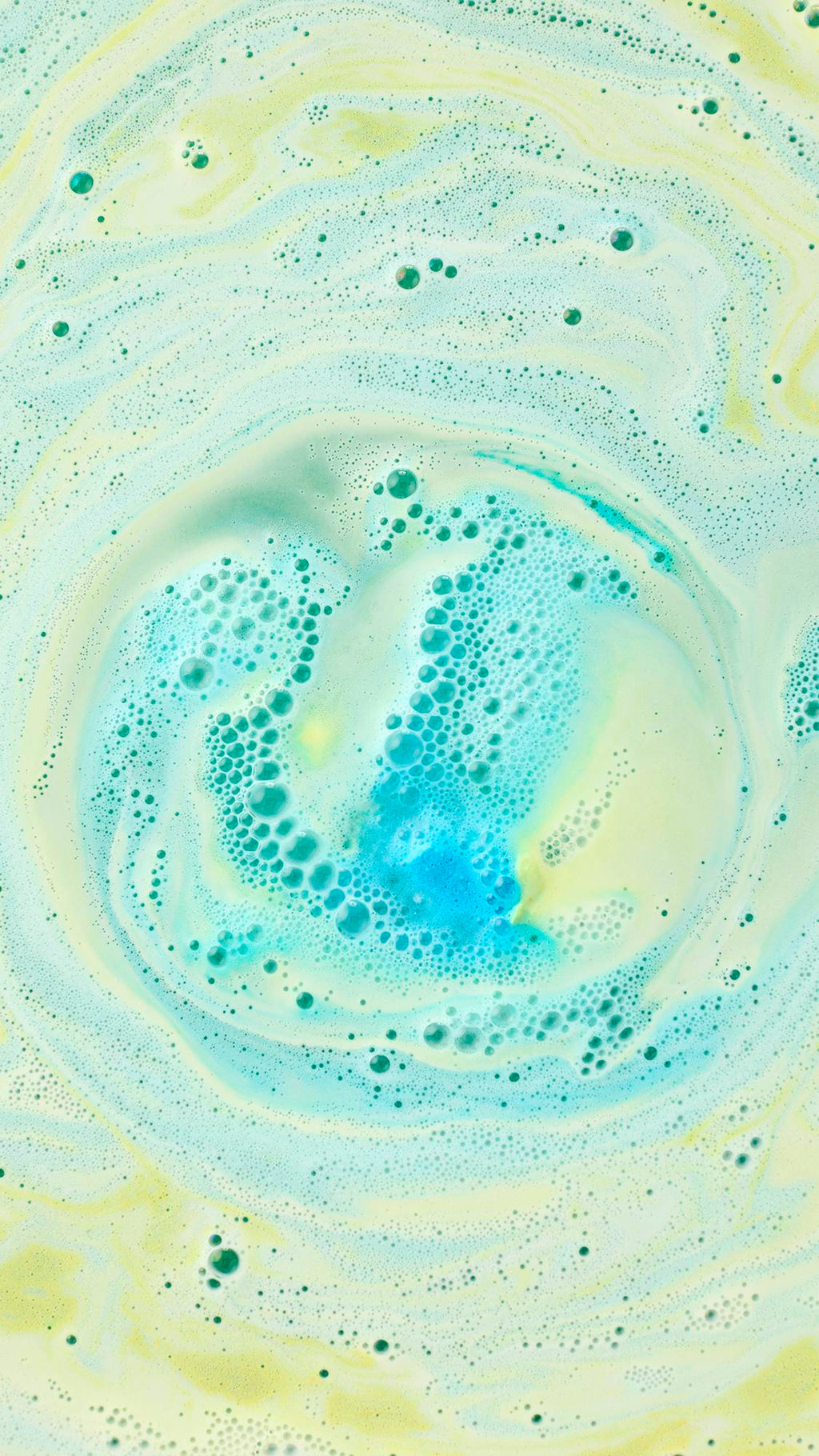 A top-down view of a swirling whirlpool of pastel green and soft yellow foam in a bath. At the center, a cluster of bright blue bubbles creates a vibrant contrast, forming a marble-like pattern across the water's surface.