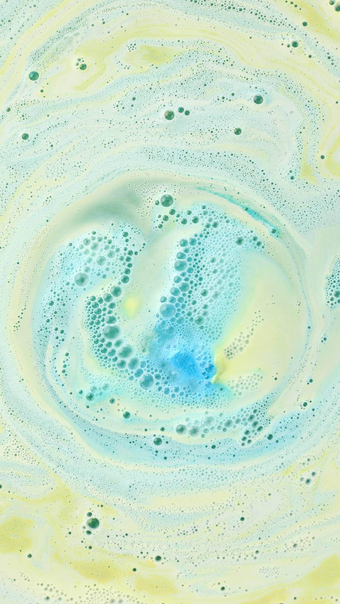A top-down view of a swirling whirlpool of pastel green and soft yellow foam in a bath. At the center, a cluster of bright blue bubbles creates a vibrant contrast, forming a marble-like pattern across the water's surface.