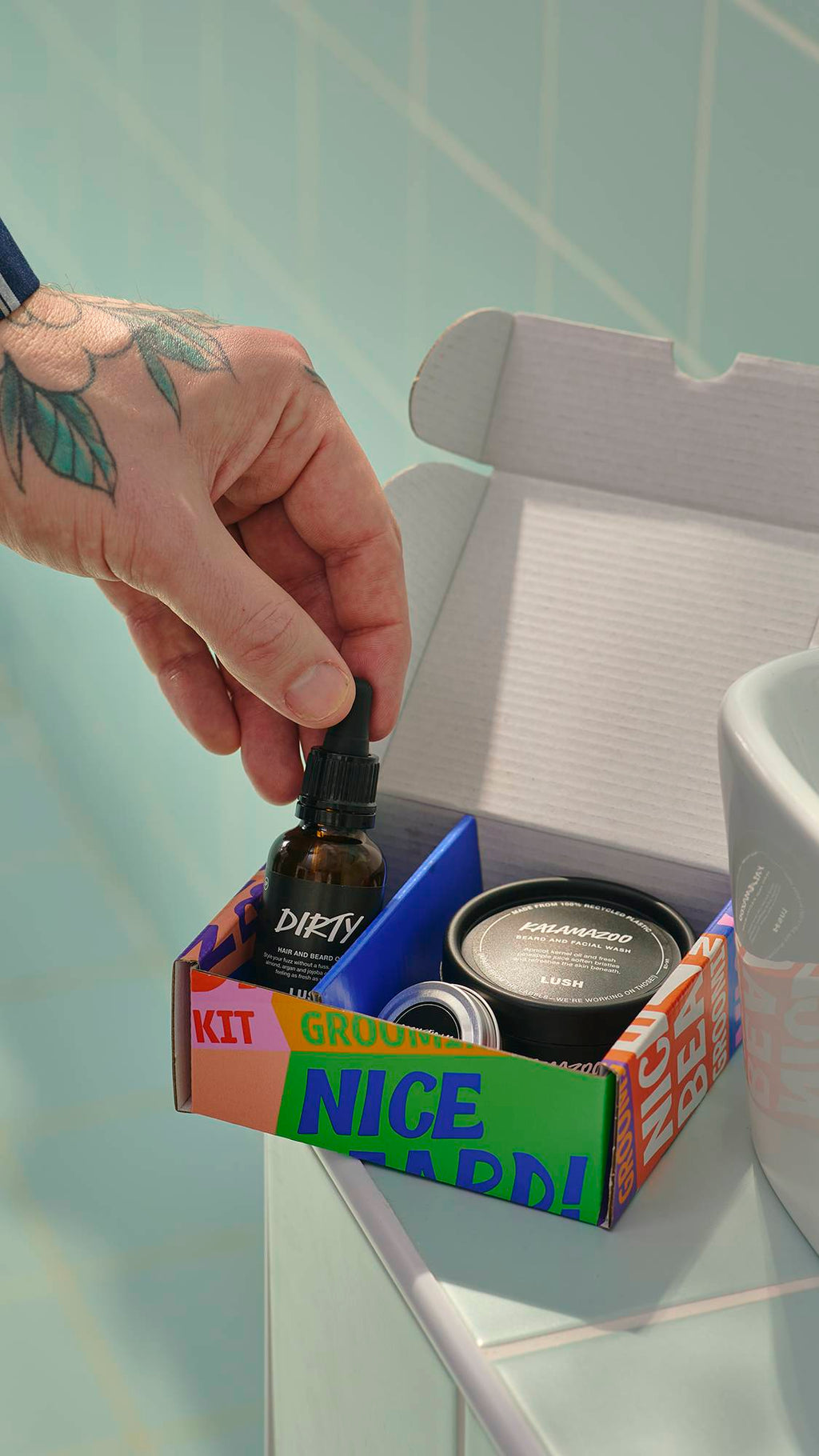 A tattooed hand reaches into an open 'Nice Beard!' grooming kit to pick up a glass bottle of Dirty hair and beard oil. The kit, which also contains a tub of face and beard wash and a tin of moisturiser, sits on the edge of a sink against blue tiles.