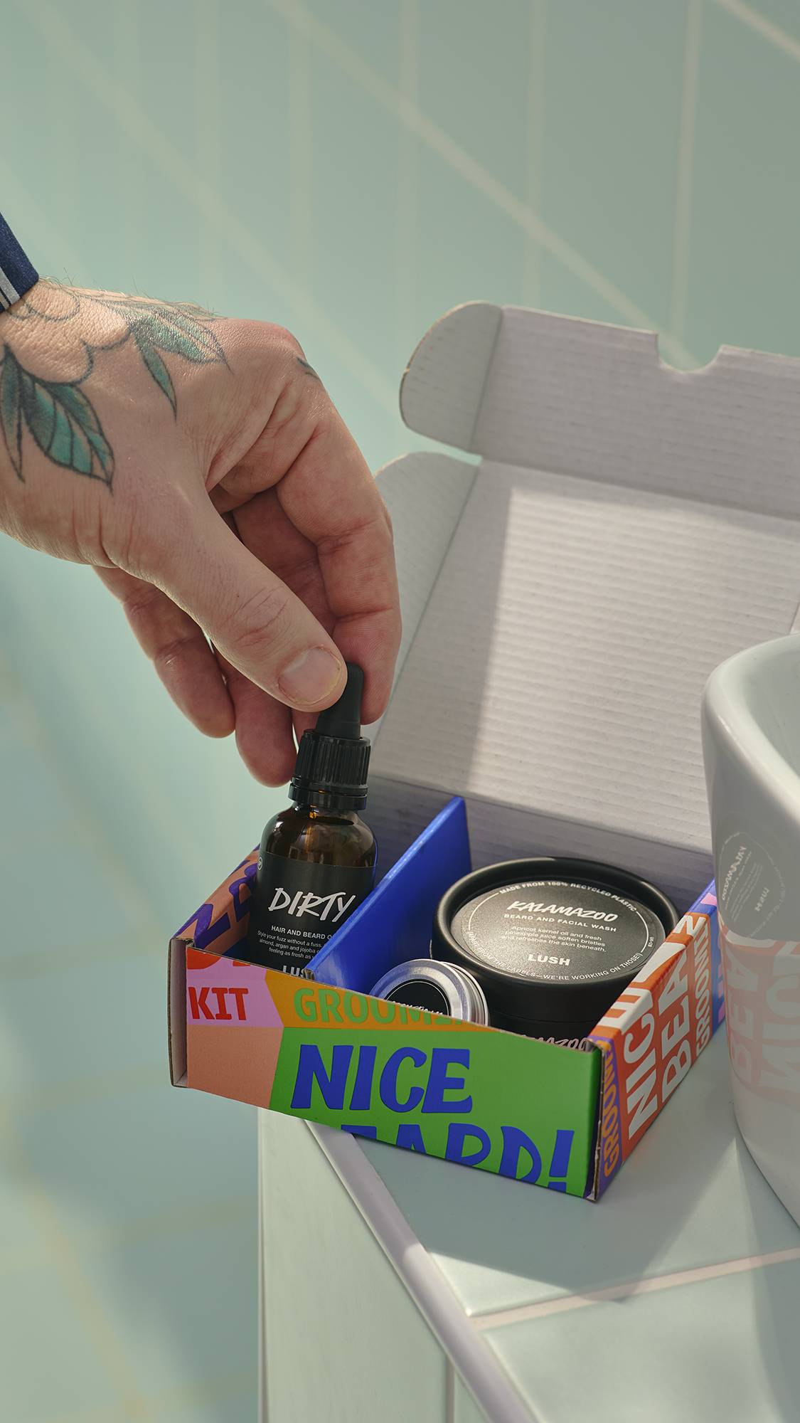 A tattooed hand reaches into an open 'Nice Beard!' grooming kit to pick up a glass bottle of Dirty hair and beard oil. The kit, which also contains a tub of face and beard wash and a tin of moisturiser, sits on the edge of a sink against blue tiles.
