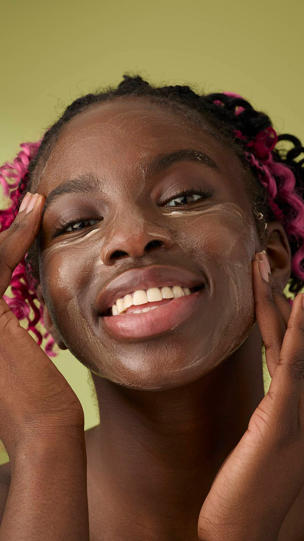 A woman with pink and black braids smiles as she applies a smooth, ivory Oat facial cleanser over her face. The thick, creamy product stands out against her skin and the solid olive-green background, emphasising its rich texture.