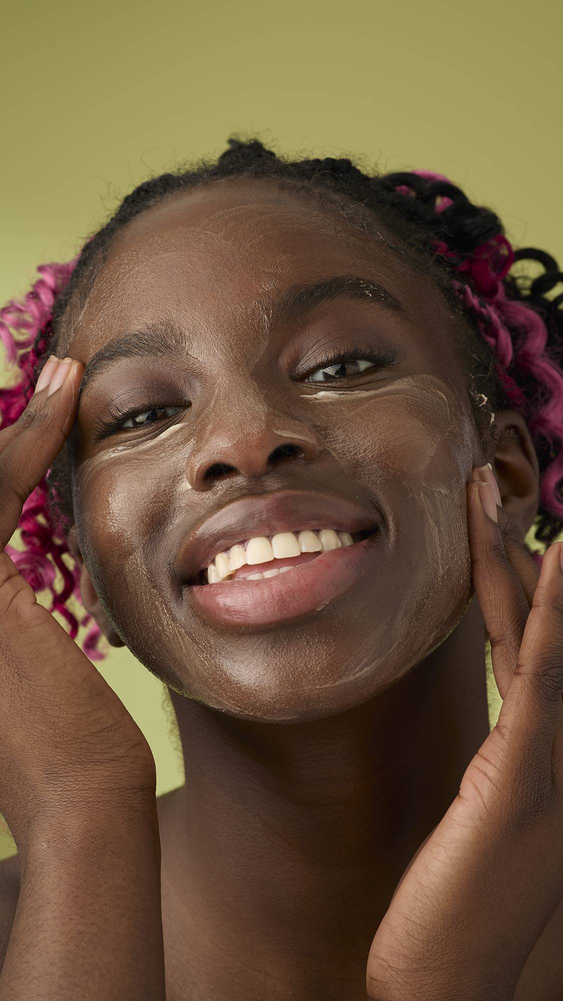 A woman with pink and black braids smiles as she applies a smooth, ivory Oat facial cleanser over her face. The thick, creamy product stands out against her skin and the solid olive-green background, emphasising its rich texture.