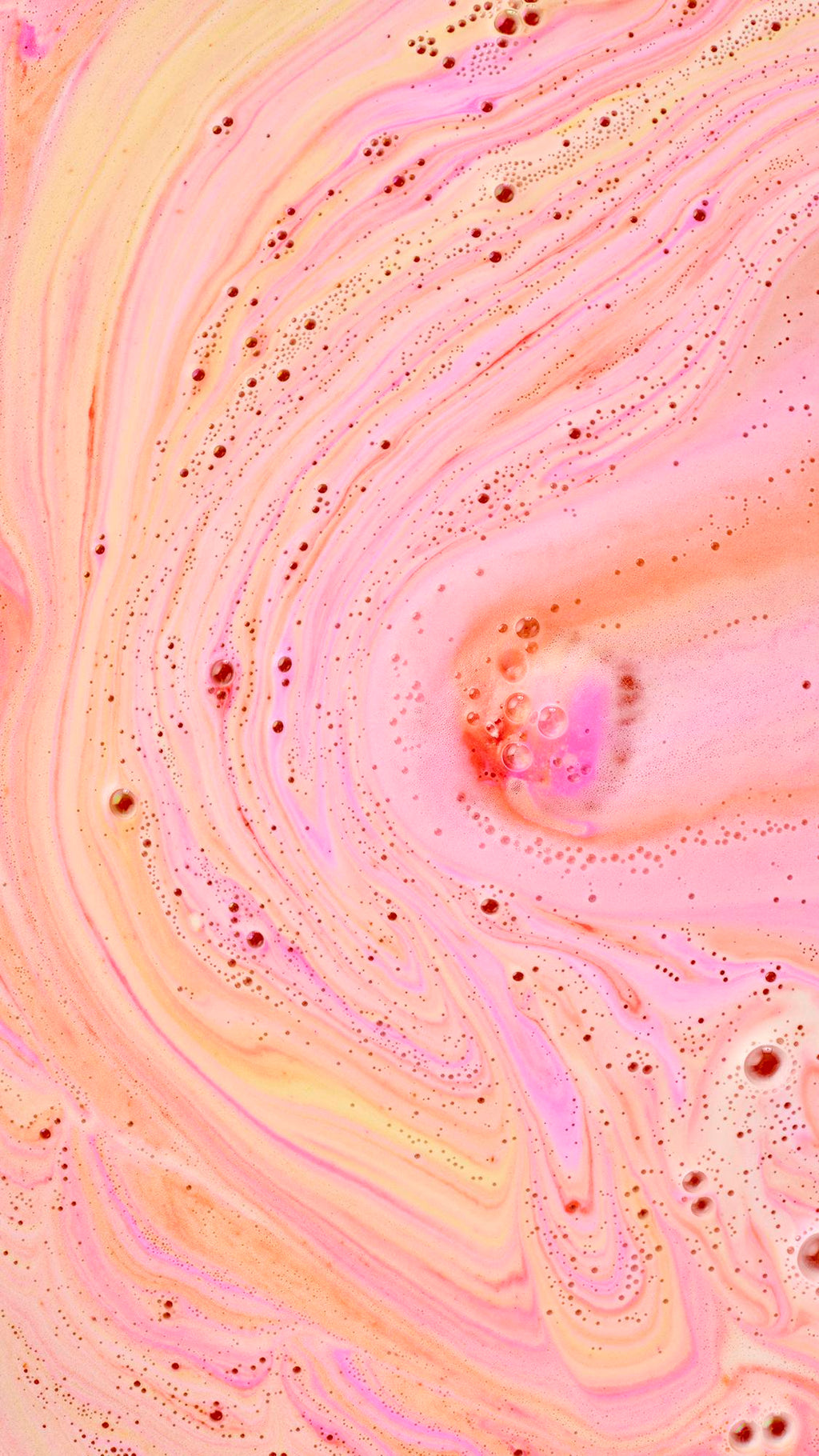 A top-down, close-up view of vibrant "bath art." Swirling, marble-like patterns of neon pink, soft orange, and pale yellow foam cover the water's surface, dotted with tiny, clear bubbles that add texture to the warm-toned display.