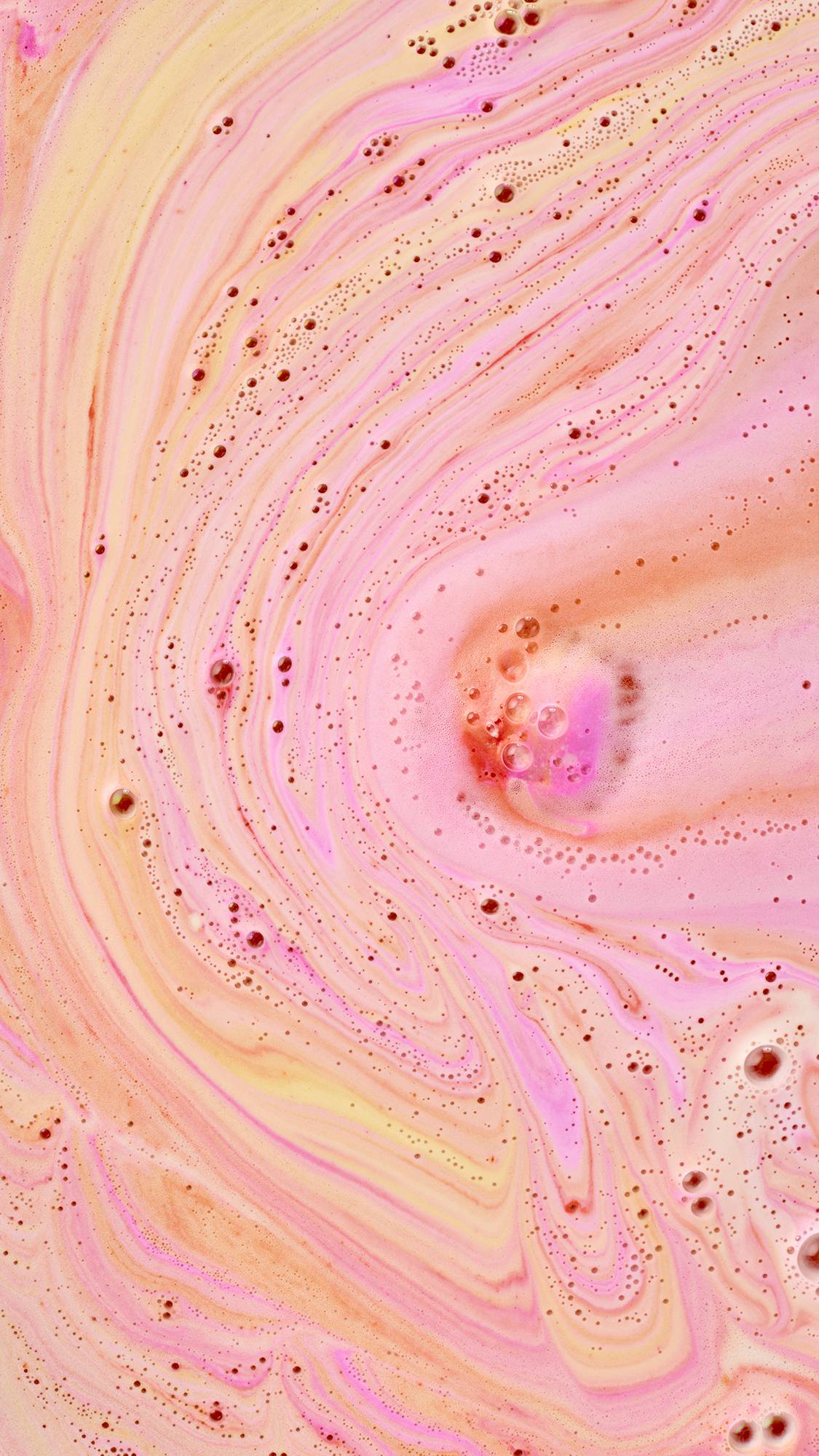 A top-down, close-up view of vibrant "bath art." Swirling, marble-like patterns of neon pink, soft orange, and pale yellow foam cover the water's surface, dotted with tiny, clear bubbles that add texture to the warm-toned display.