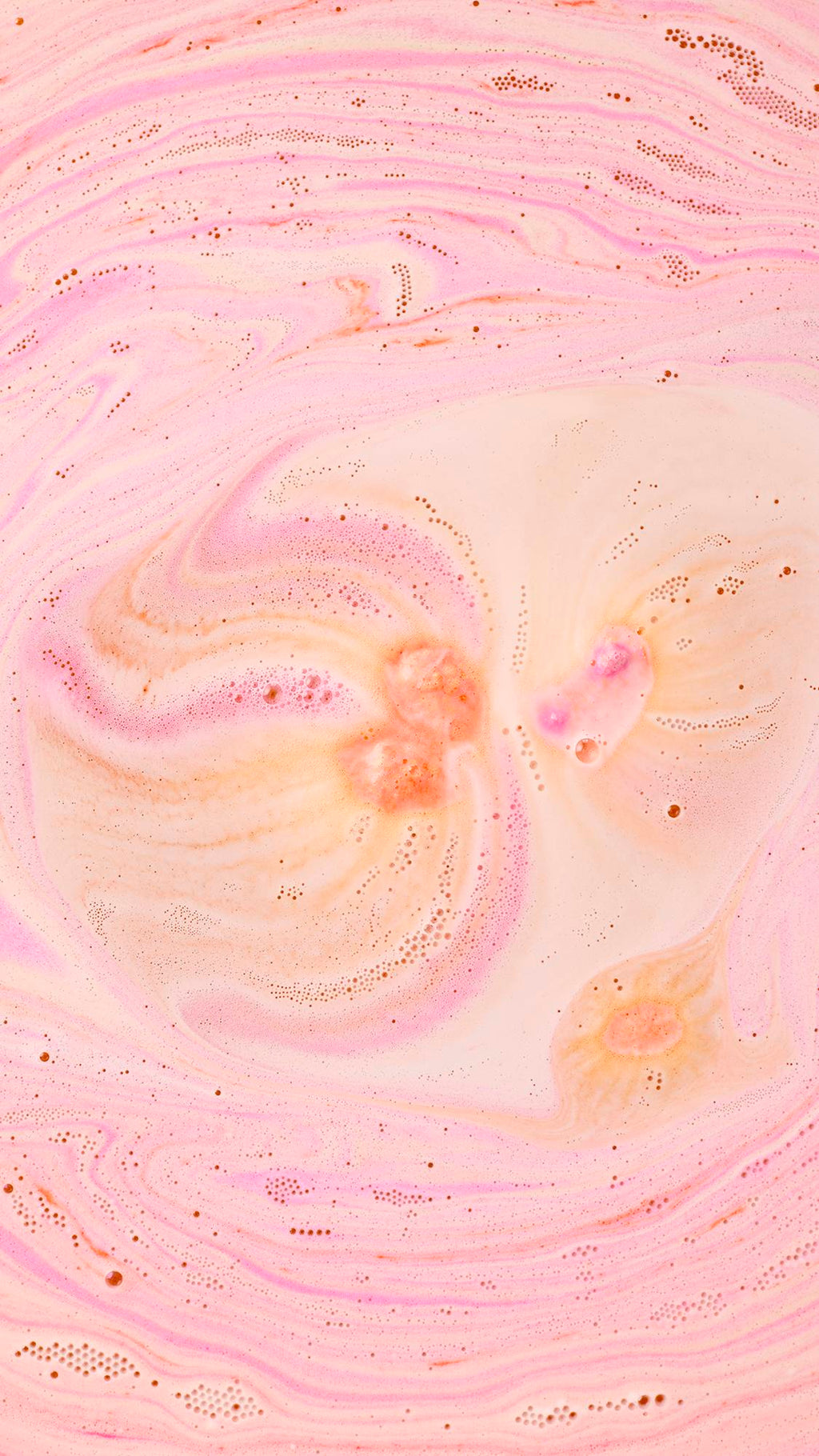 An aerial view of bathwater filled with swirling patterns of peach, pink, and cream. Delicate trails of tiny bubbles and frothy foam create an intricate, marbled texture across the surface, spiralling outward from central points of colour.