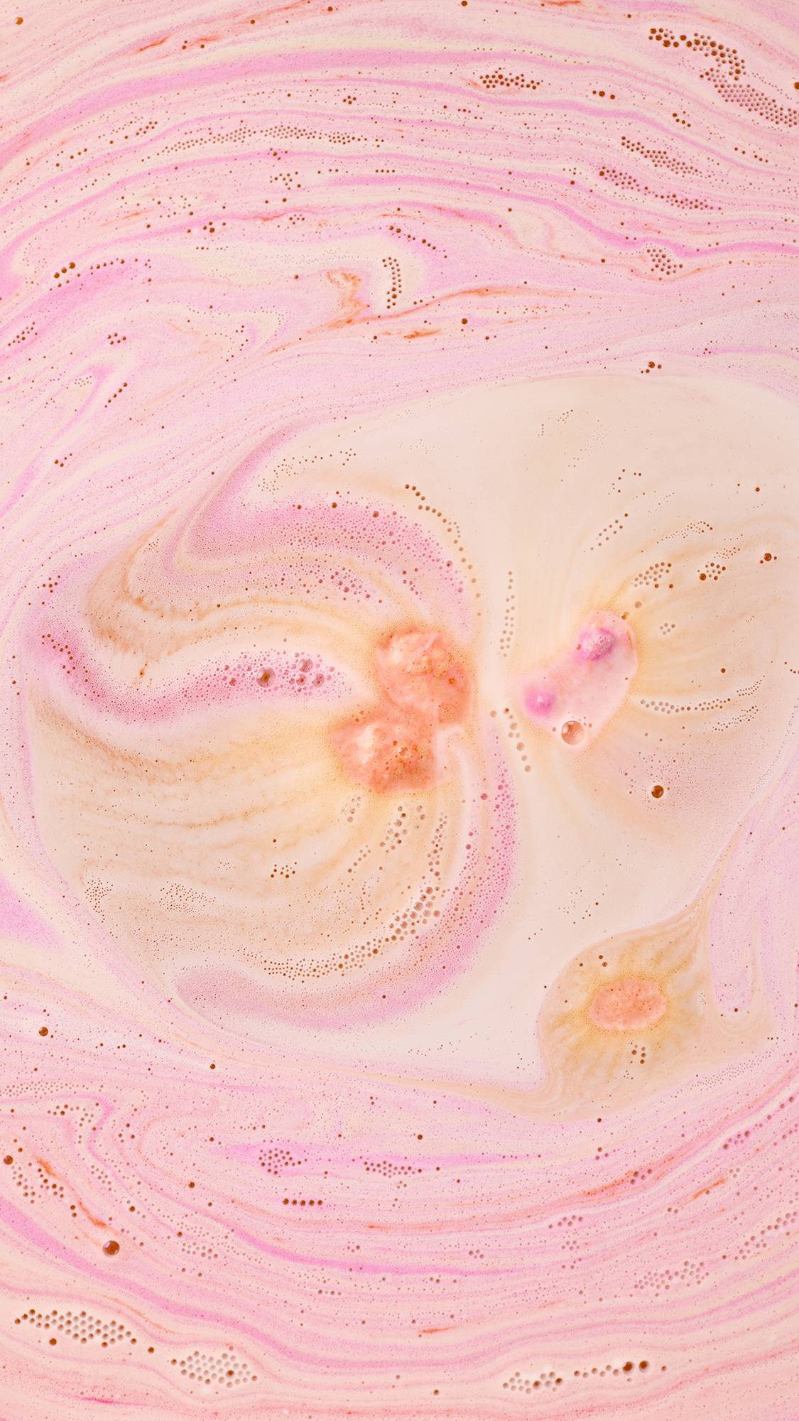 An aerial view of bathwater filled with swirling patterns of peach, pink, and cream. Delicate trails of tiny bubbles and frothy foam create an intricate, marbled texture across the surface, spiralling outward from central points of colour.