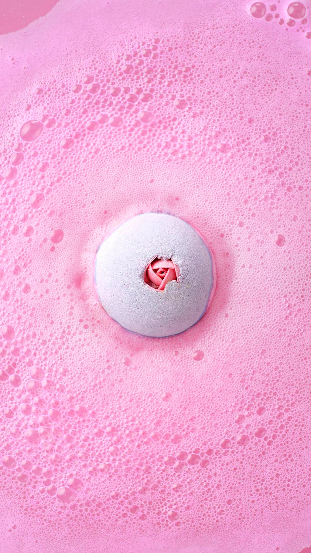 Top-down view of the Sex Bomb Bath Bomb dissolving in a sea of vibrant, frothy pink bubbles, with its signature rosebud detail visible in the center.