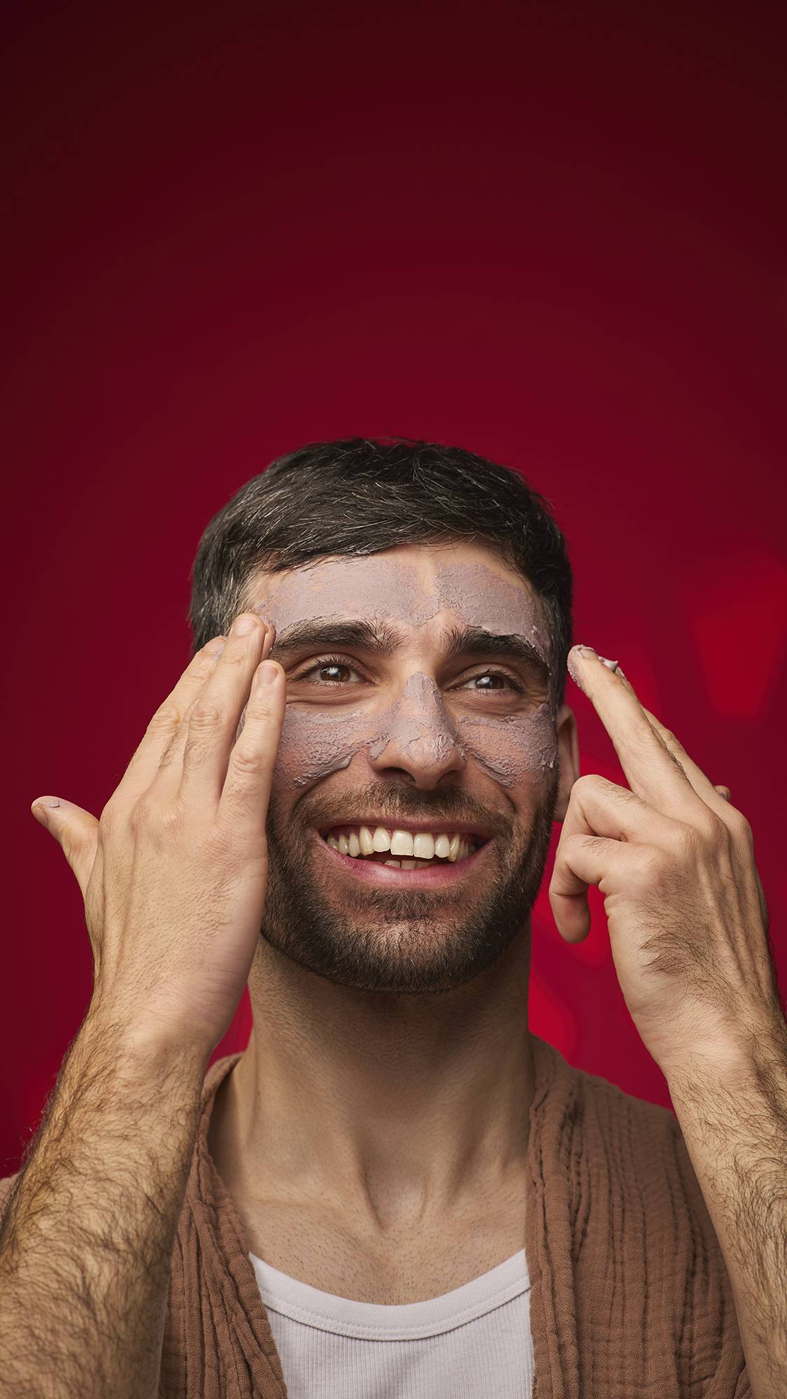 A smiling person applies the mauve-toned Sipak face mask to their forehead and nose. They wear a brown textured robe against a solid deep red background.