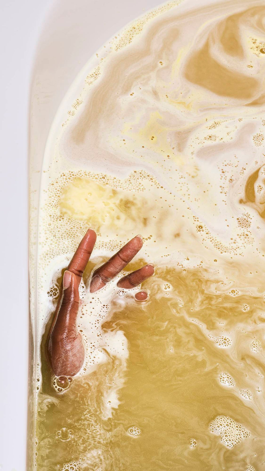 A close-up of a model's hands submerged in a golden bath. Thick, creamy yellow foam swirls across the water's surface as a bath bomb dissolves, creating a marbled pattern of light and dark gold.