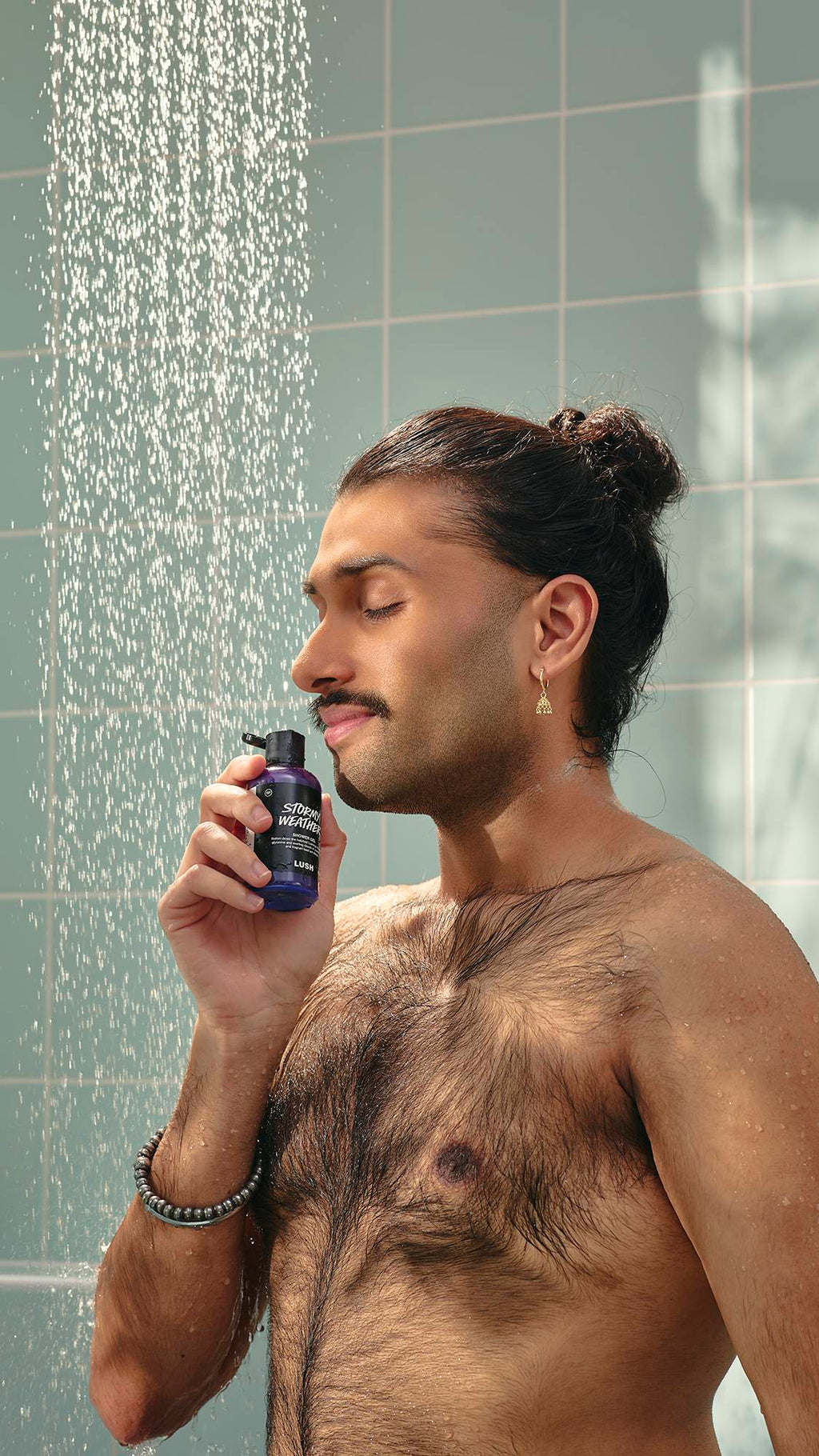 A model with dark hair in a bun and a moustache stands in a shower, holding a bottle of purple "Stormy Weather" shower gel to their nose. They have their eyes closed with a peaceful expression as water falls around them against pale green tiles.