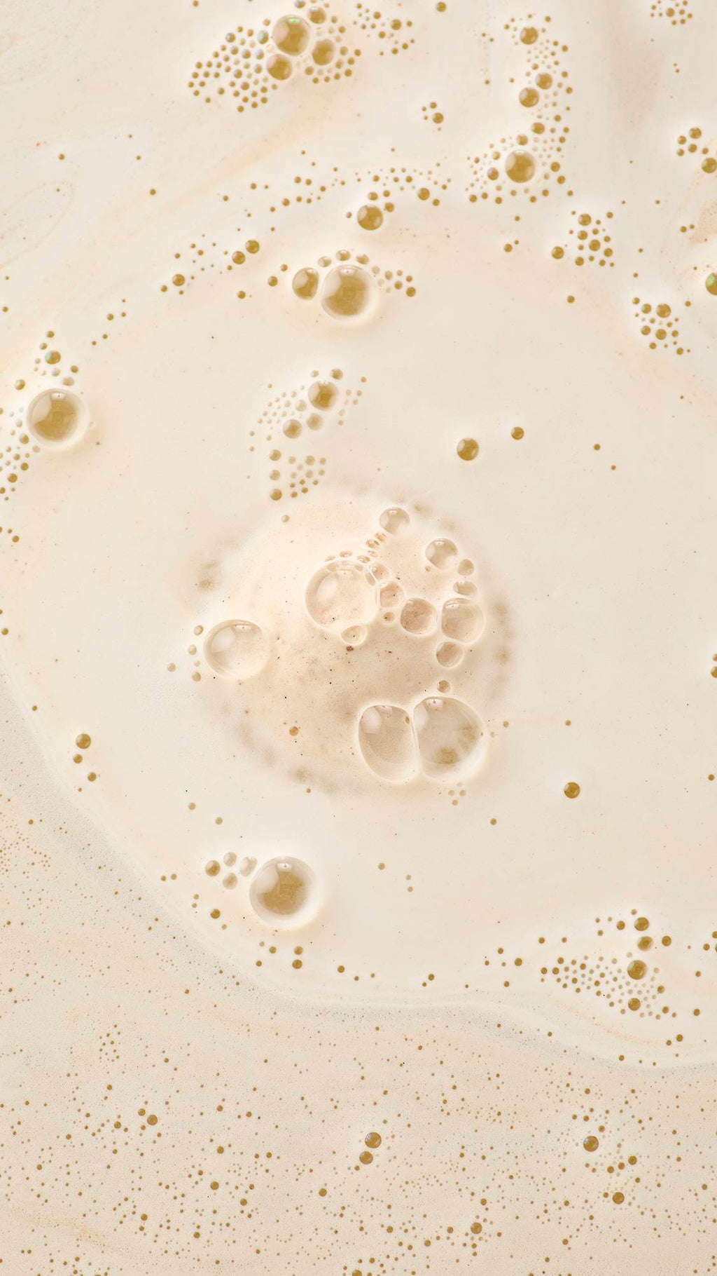 An aerial, abstract view of creamy bathwater. Fine, frothy bubbles and larger translucent suds cluster and swirl across the surface, creating a marbled and textured effect of velvety foam.