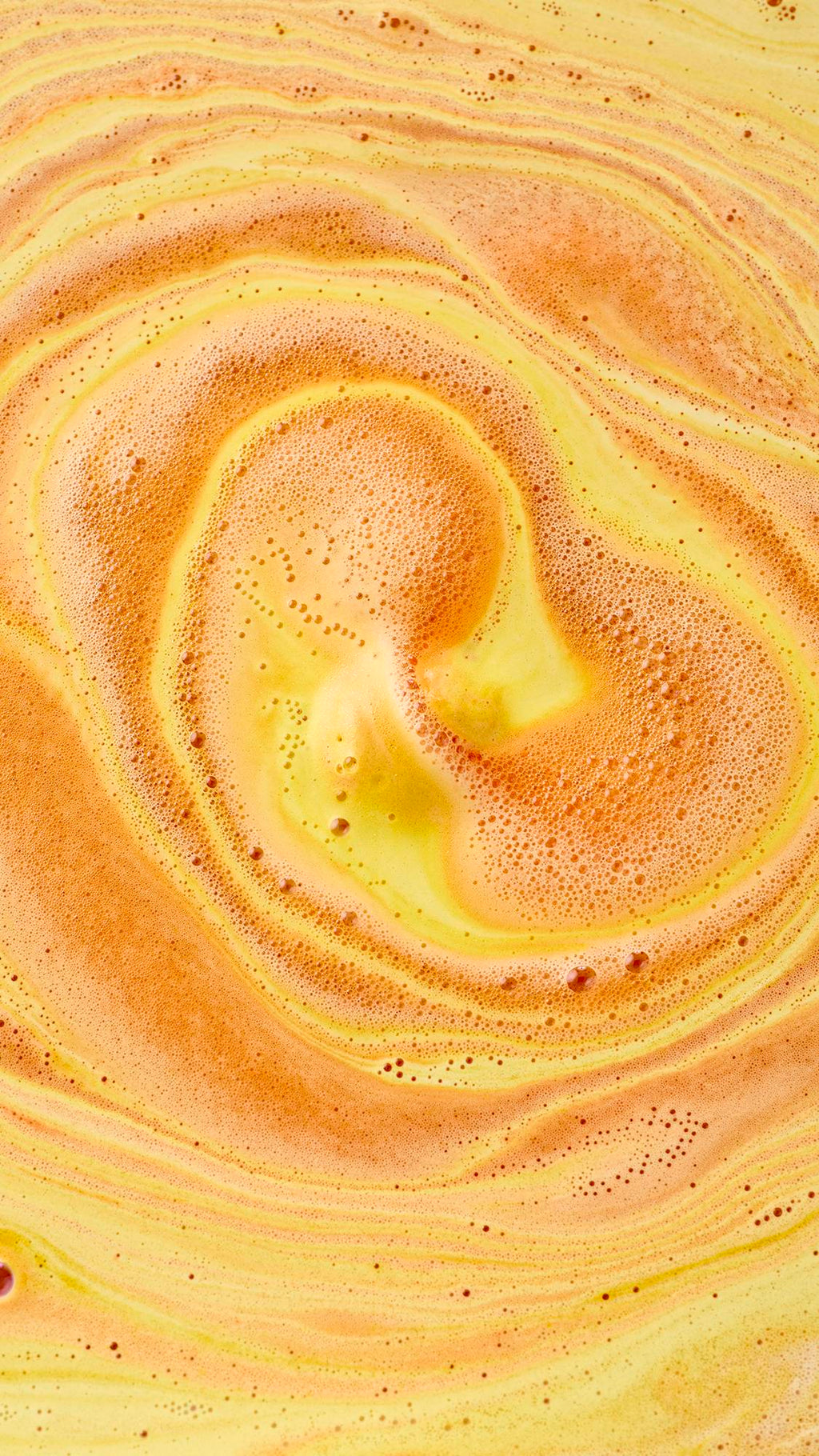 AN aerial, abstract view of swirling bath art created by the "Sweet As Honey" bath bomb. Vibrant streaks of amber-orange foam and pale yellow liquid spiral together, creating a marbled, creamy texture with tiny bubbles across the surface.
