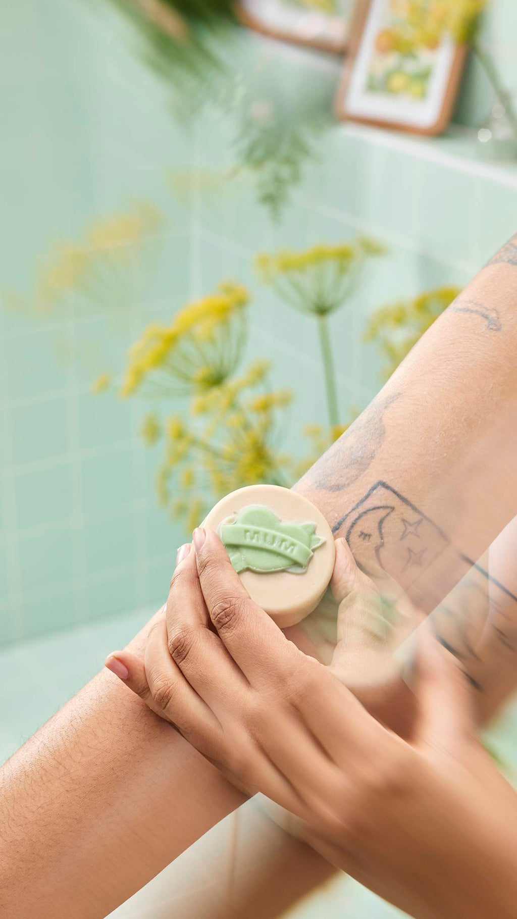 Hands massage a cream "Symbol of Love" oil bar with a green "MUM" banner onto a tattooed arm. Yellow flowers frame the scene against a mint green tiled background.