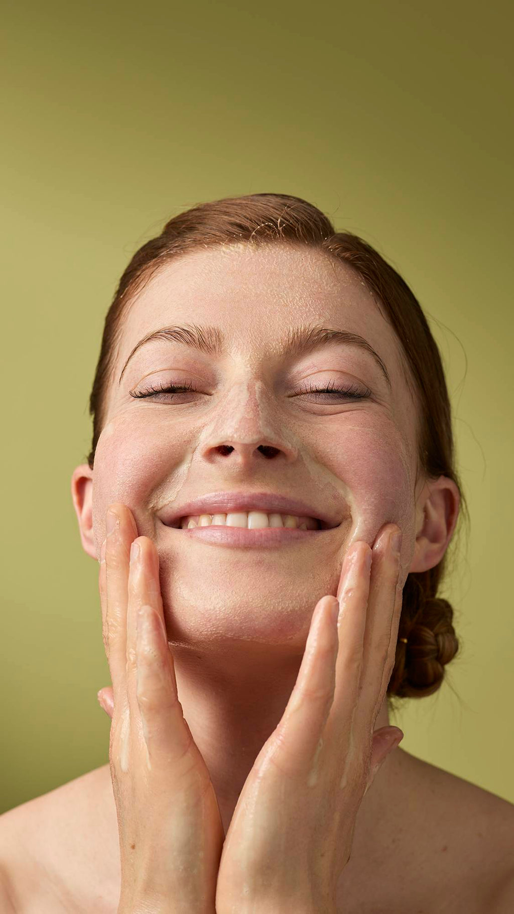 A person with their eyes closed and a gentle smile massages a foamy cleanser into their face. Their wet hands are covered in light soap suds, and they stand against a solid olive-green background.