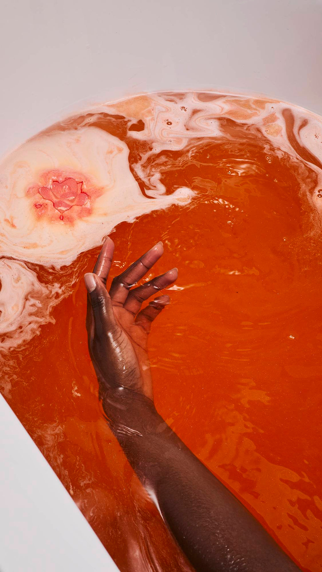 The image shows the "Comforter Rose" bath bomb dissolving in bathwater, creating a large island of thick white foam. A model's hand reaches into the deep orange, shimmering water toward the foam, which features a pink floral pattern in its centre.