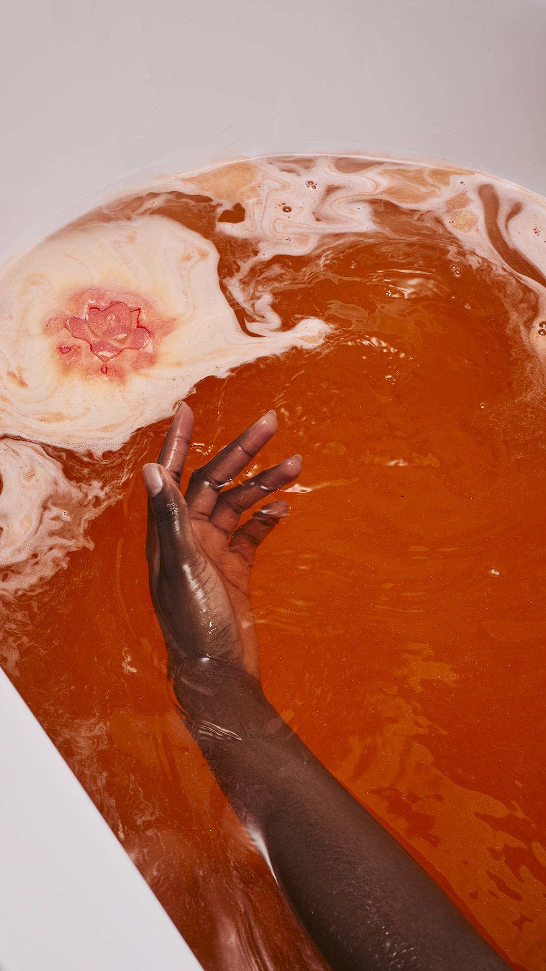 The image shows the "Comforter Rose" bath bomb dissolving in bathwater, creating a large island of thick white foam. A model's hand reaches into the deep orange, shimmering water toward the foam, which features a pink floral pattern in its centre.