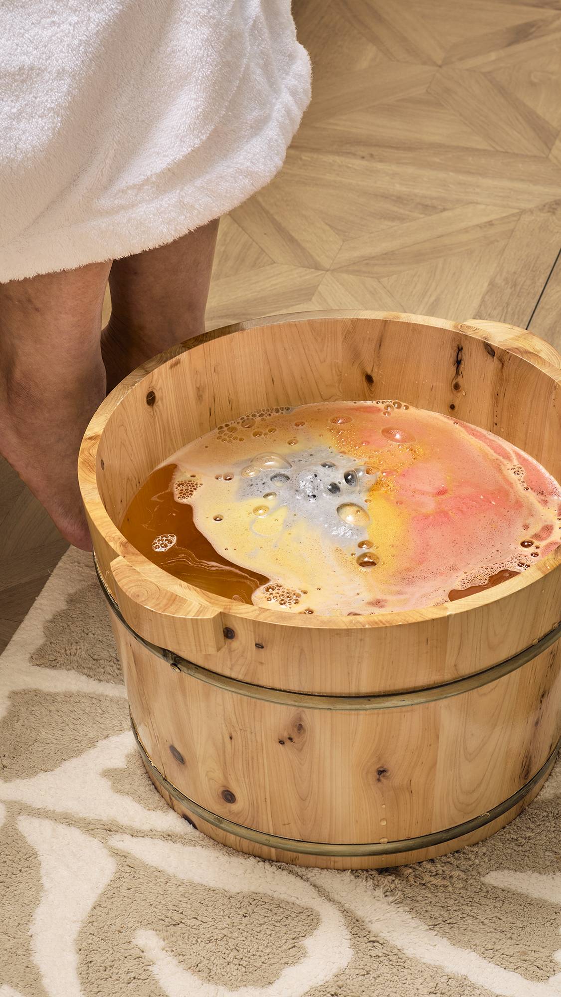 The image shows the "Volcano" foot soak as it dissolves in a wooden foot tub, turning the water a deep golden-orange. Swirls of grey, yellow, and pink foam spread across the surface, creating a colourful, bubbling display as it fizzes away.