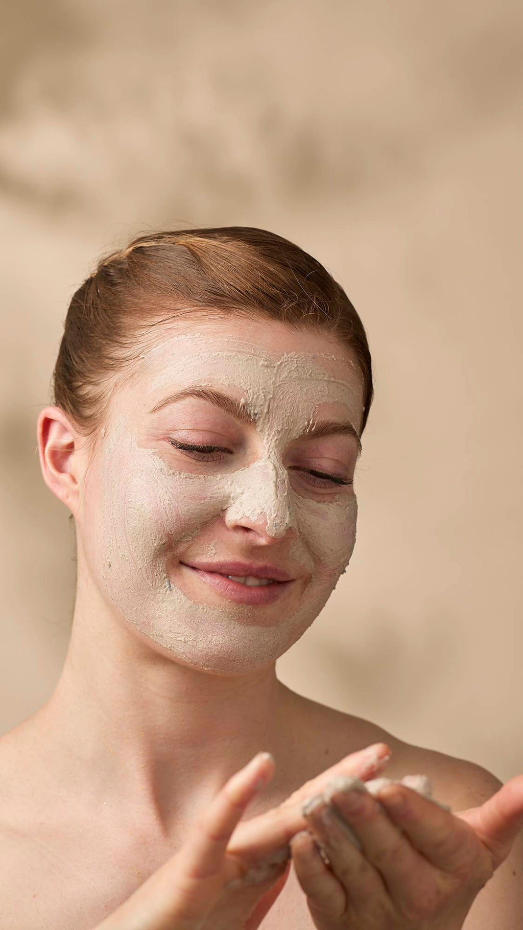 A model with their hair tied back looks down and smiles as they apply a thick, beige "Tiny Musicians Of The Forest" facial cleanser. The creamy, textured mask covers most of their face as they rub more of the product between their hands.