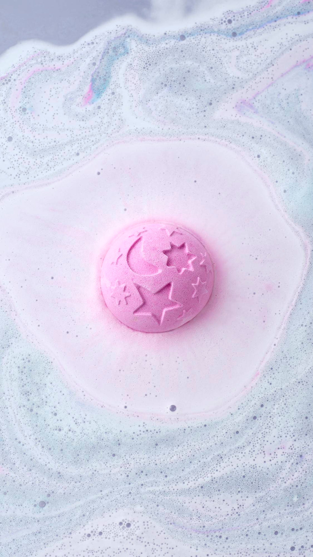 A top-down view shows the Twilight Bath Bomb dissolving, creating a soft pink halo in the water. The lavender sphere, featuring embossed celestial designs, floats amidst a sea of frothy white and light blue foam.